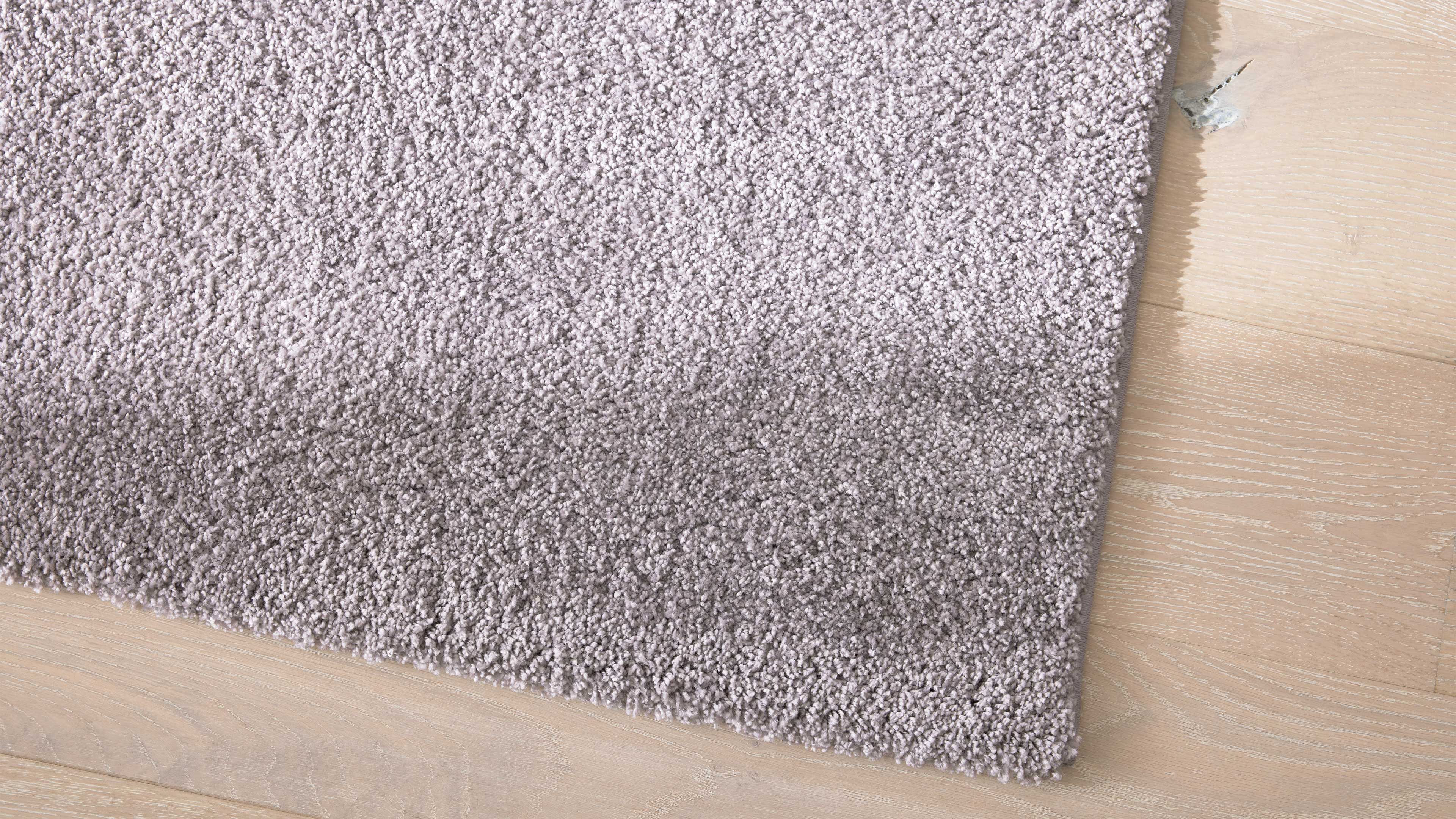 Hellgraue, hochflorige Teppichecke liegt auf hellen Holzdielen mit sichtbarer Maserung und einer kleinen Macke im Holz.