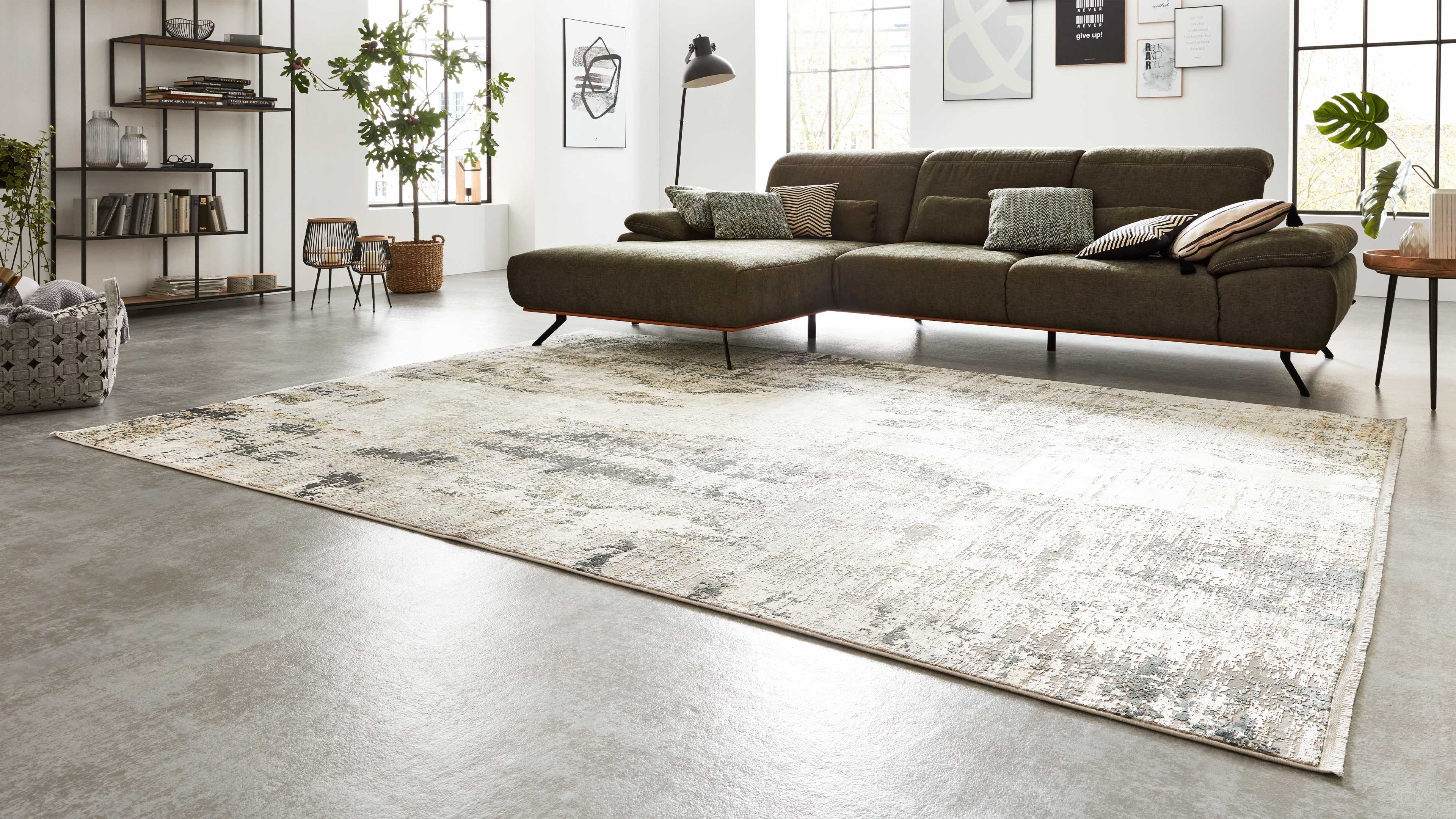 Olivgrünes Ecksofa mit Chaiselongue, Kissen und Stehlampe, davor großer hell gemusterter Teppich, Regal und Pflanzen im Raum.