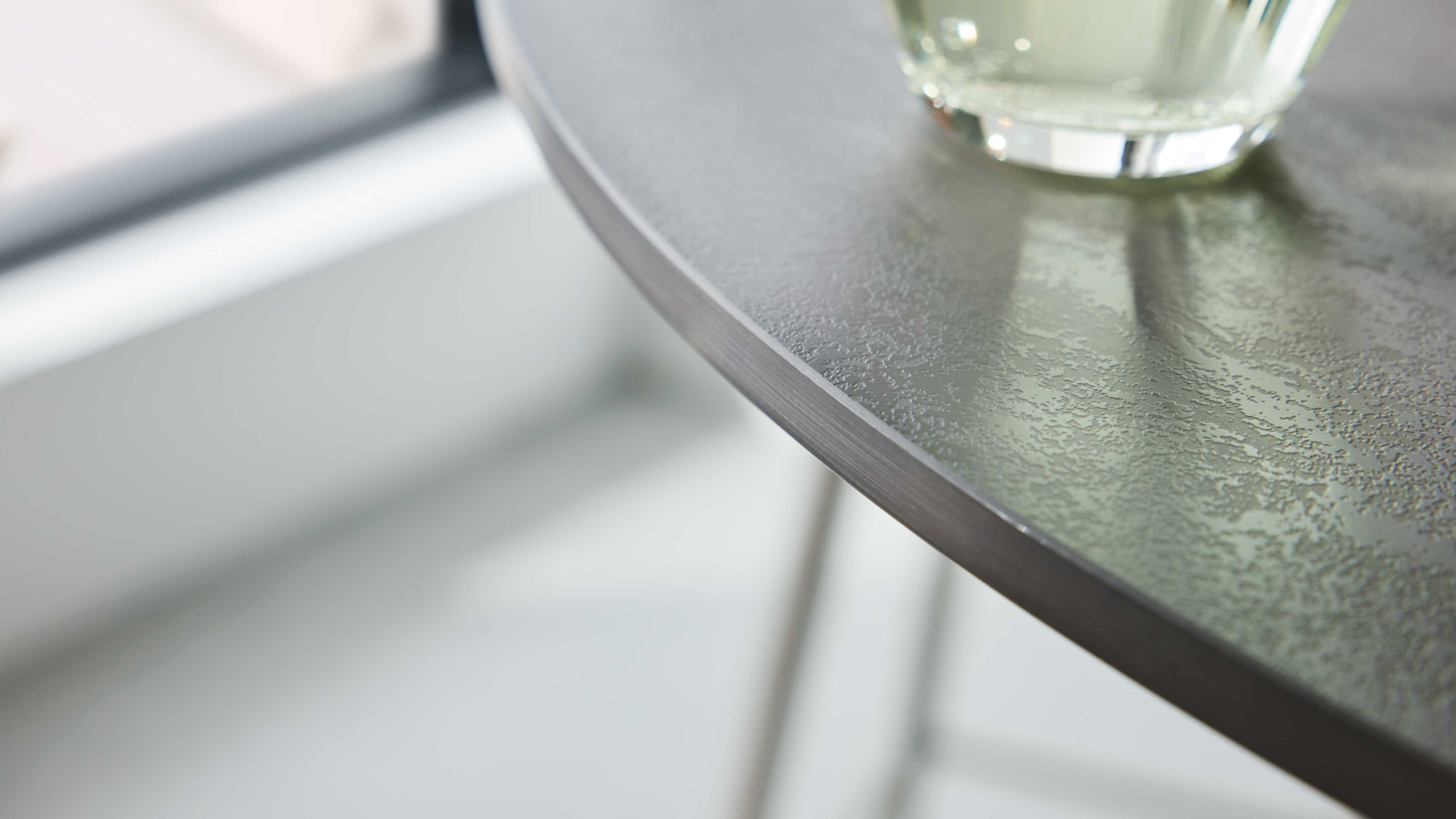 Runder Tisch mit dunkelgrauer, leicht strukturierter Oberfläche und sichtbarer Kante, rechts Teil eines Glases mit Reflexion.