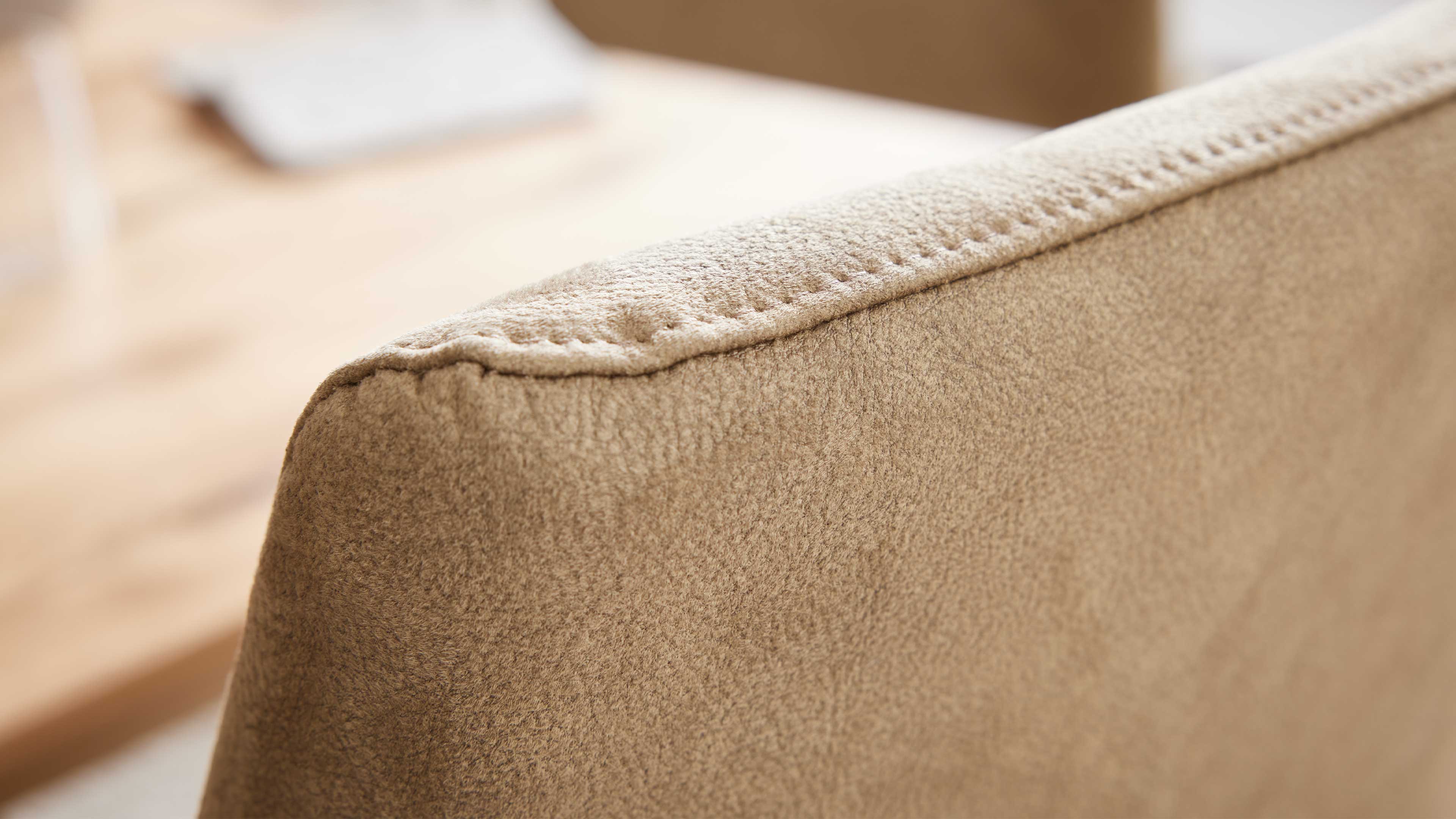 Nahaufnahme der Rückenlehne eines beige bezogenen Polstermöbels mit sichtbarer Kante und Naht, unscharfer Holztisch im Hintergrund.