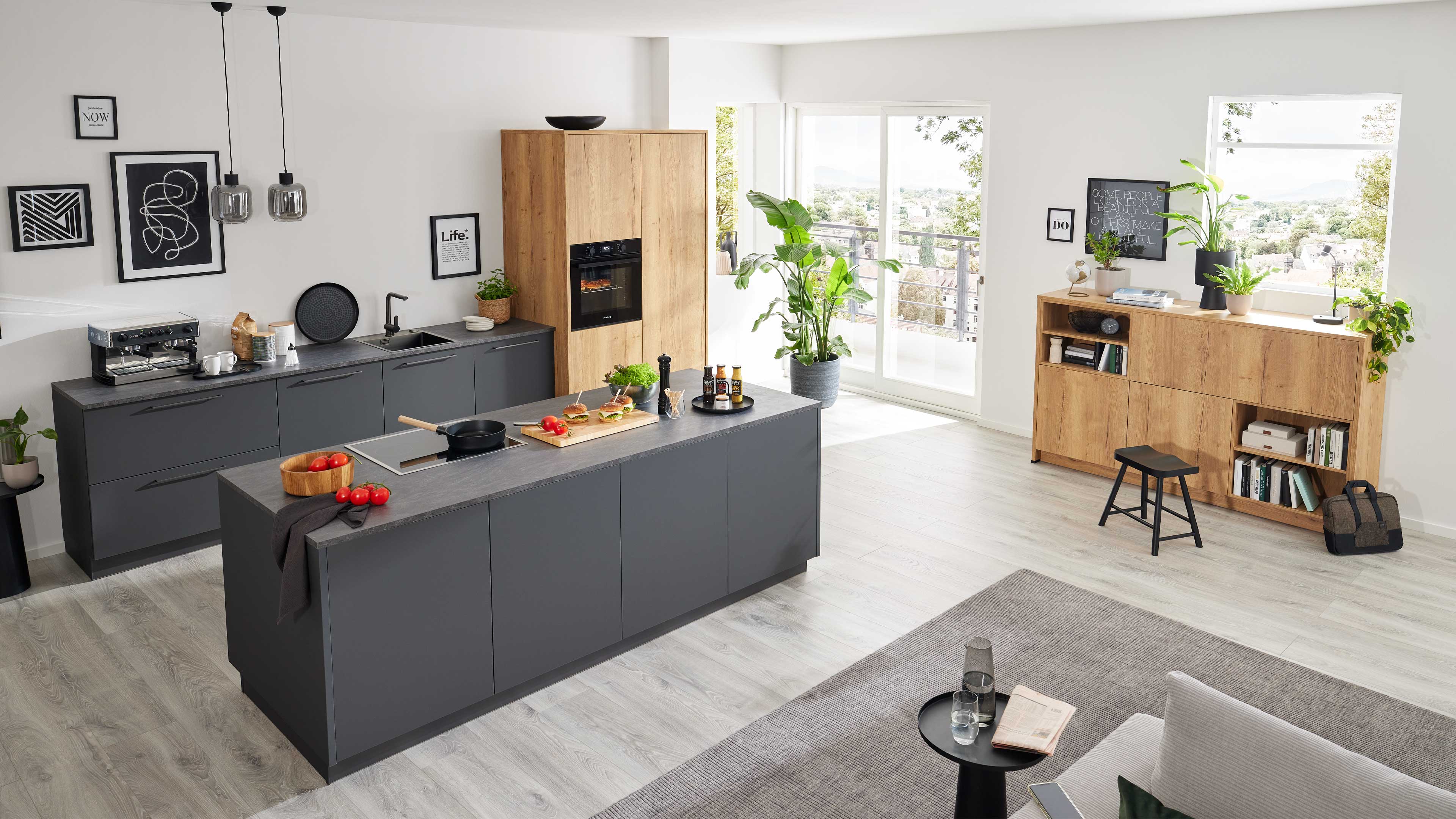 Große dunkelgraue Kücheninsel mit Kochfeld und Schneidebrett, dahinter graue Unterschränke, Eiche-Hochschrank, Sideboard und Pflanzen.