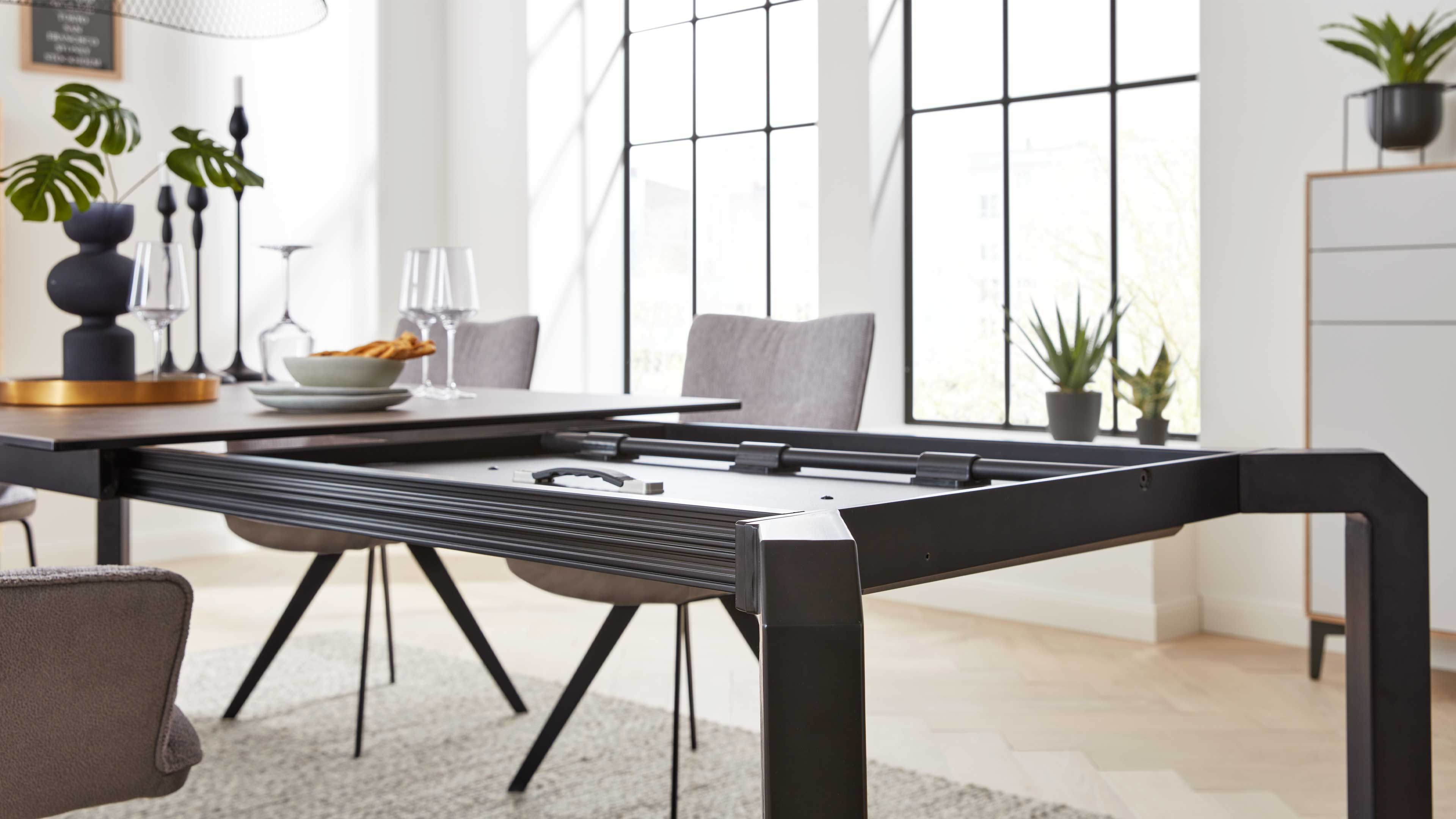 Ausziehbarer Tisch mit schwarzem Metallgestell und sichtbarer Schiene, dazu graue Stühle, Gläser und Pflanzen.