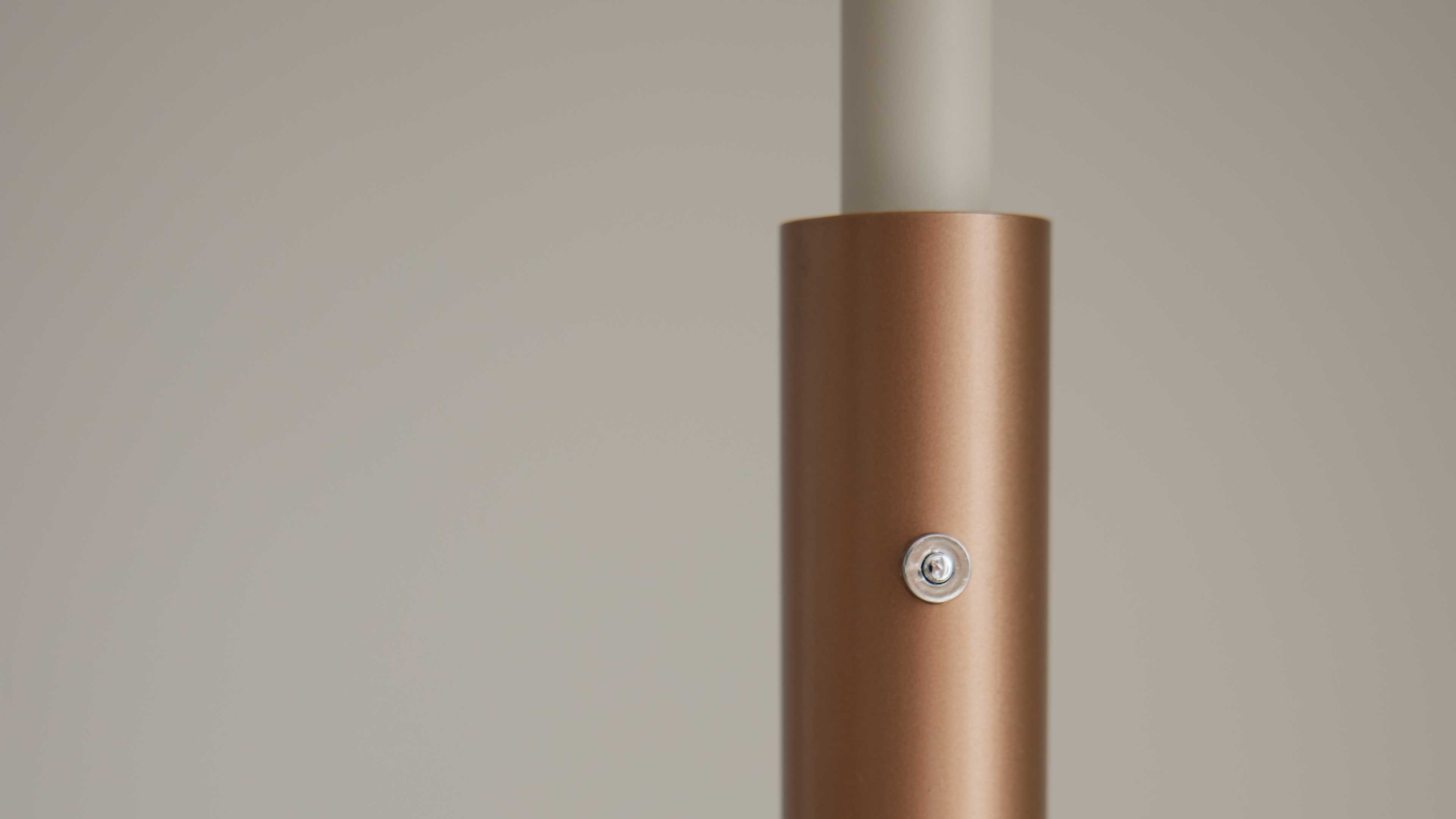 Vertikal stehendes, kupferfarbenes Metallrohr mit rundem silbernen Befestigungsdetail und einem weißen Innenrohr oben vor heller Wand.