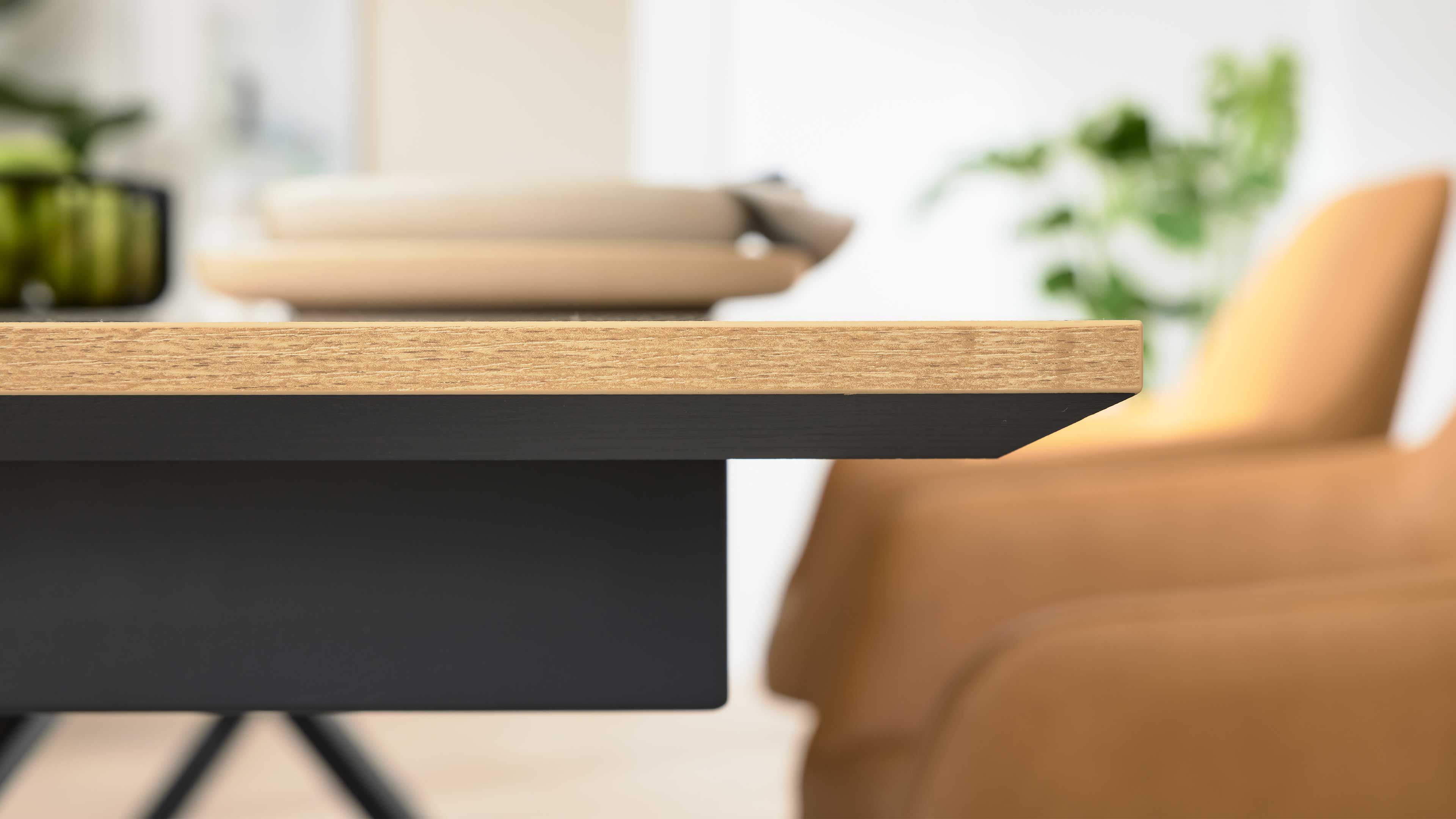 Nahaufnahme einer hellen Holz-Tischplatte mit schwarzem Unterbau, im Hintergrund unscharfer ockerfarbener Stuhl, Teller und Pflanze.