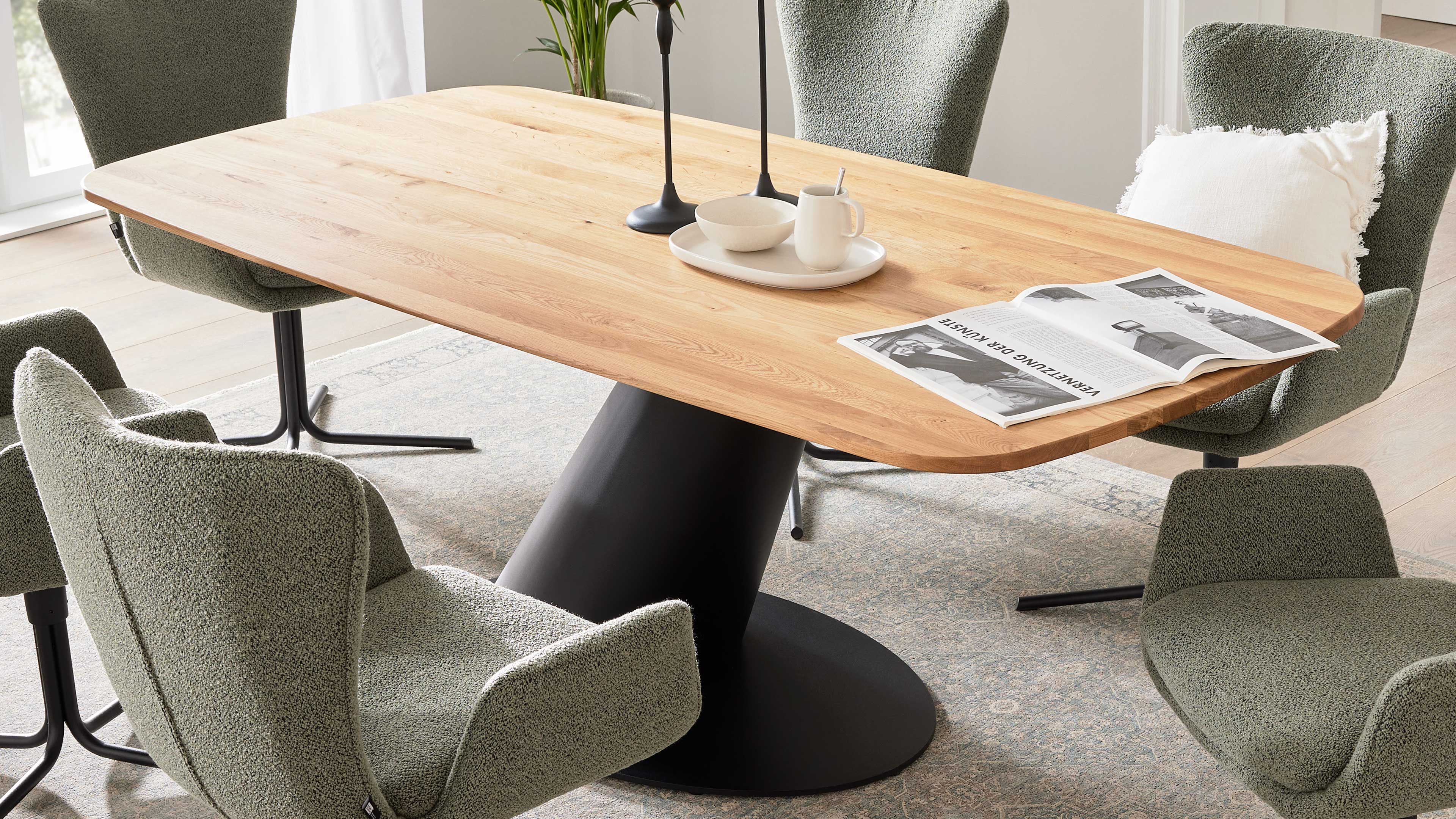 Hölzerner Tisch mit abgerundeten Kanten auf schwarzem zentralem Fuß, umgeben von fünf grünen, gepolsterten Stühlen.