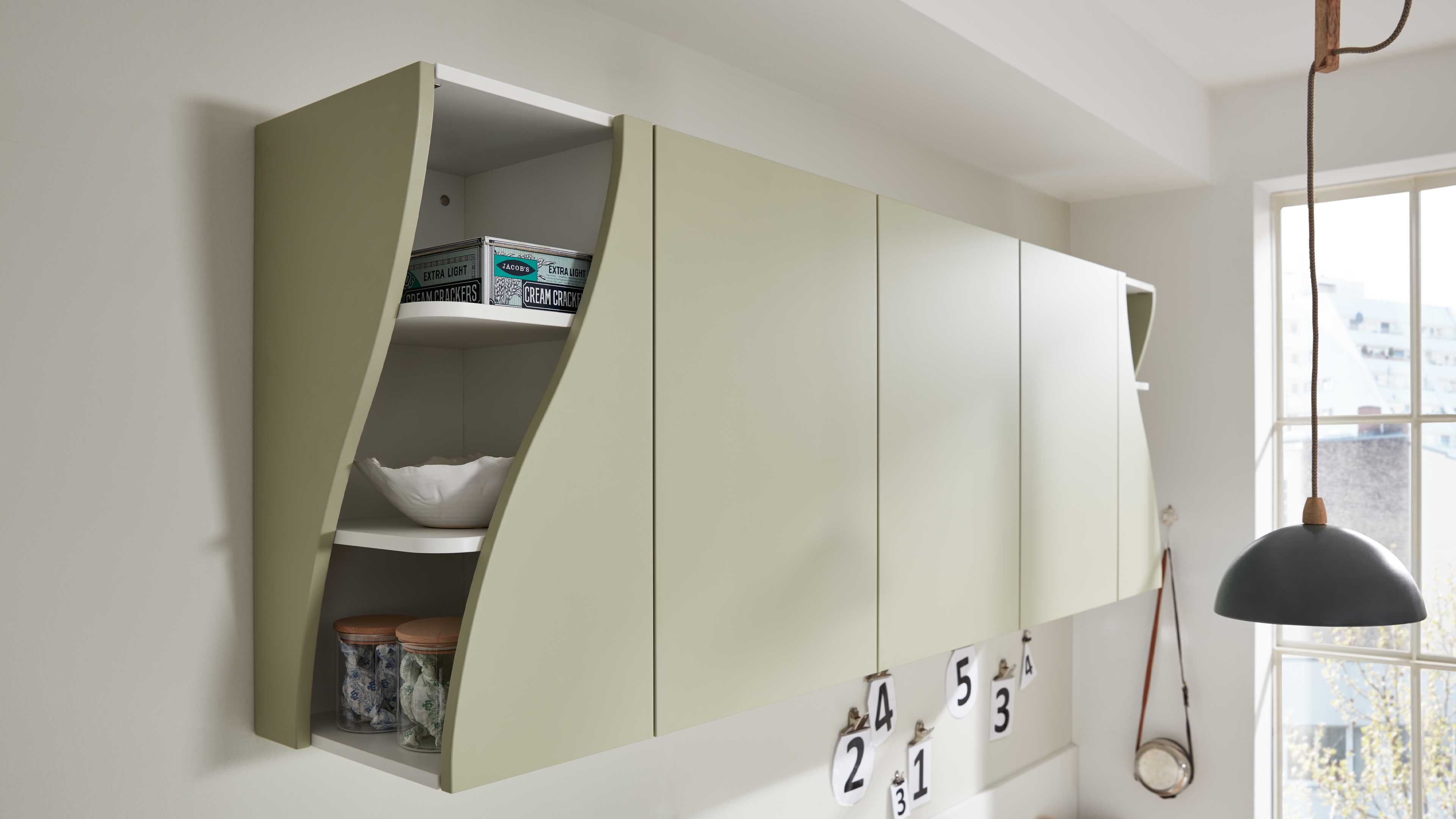 Wandhängender Küchenoberschrank in matter Hellgrün mit grifflosen Türen und offenen, geschwungenen Regalen, bestückt mit Schalen und Gläsern.