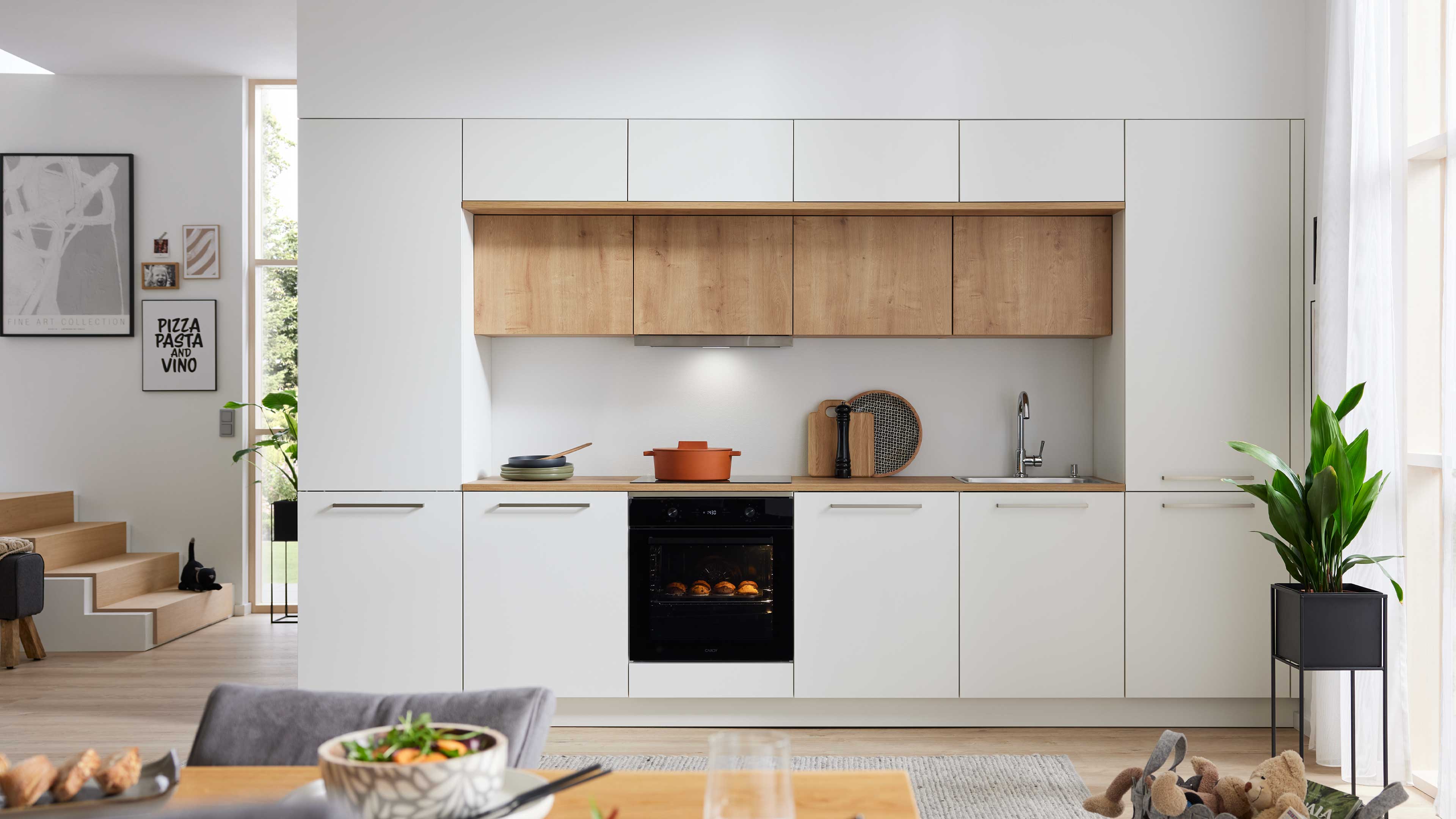 Küchenfront mit weißen Unterschränken und Holz-Oberschränken, schwarzem Einbauofen, Kochfeld, Spüle und Topf auf Arbeitsplatte.