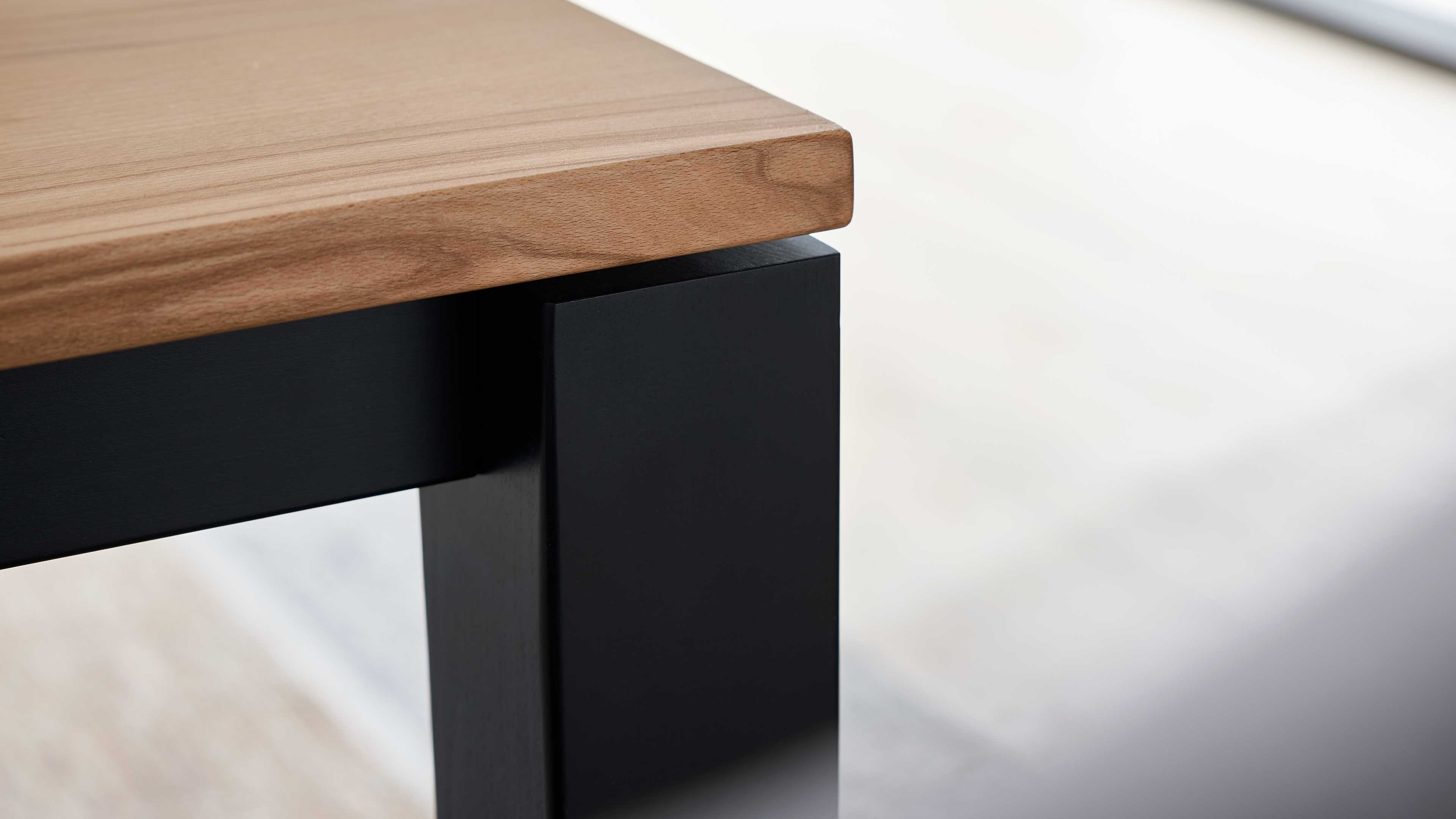 Ecke einer Tischplatte aus hellem Holz mit sichtbarer Maserung auf schwarzem, rechteckigem Tischbein.