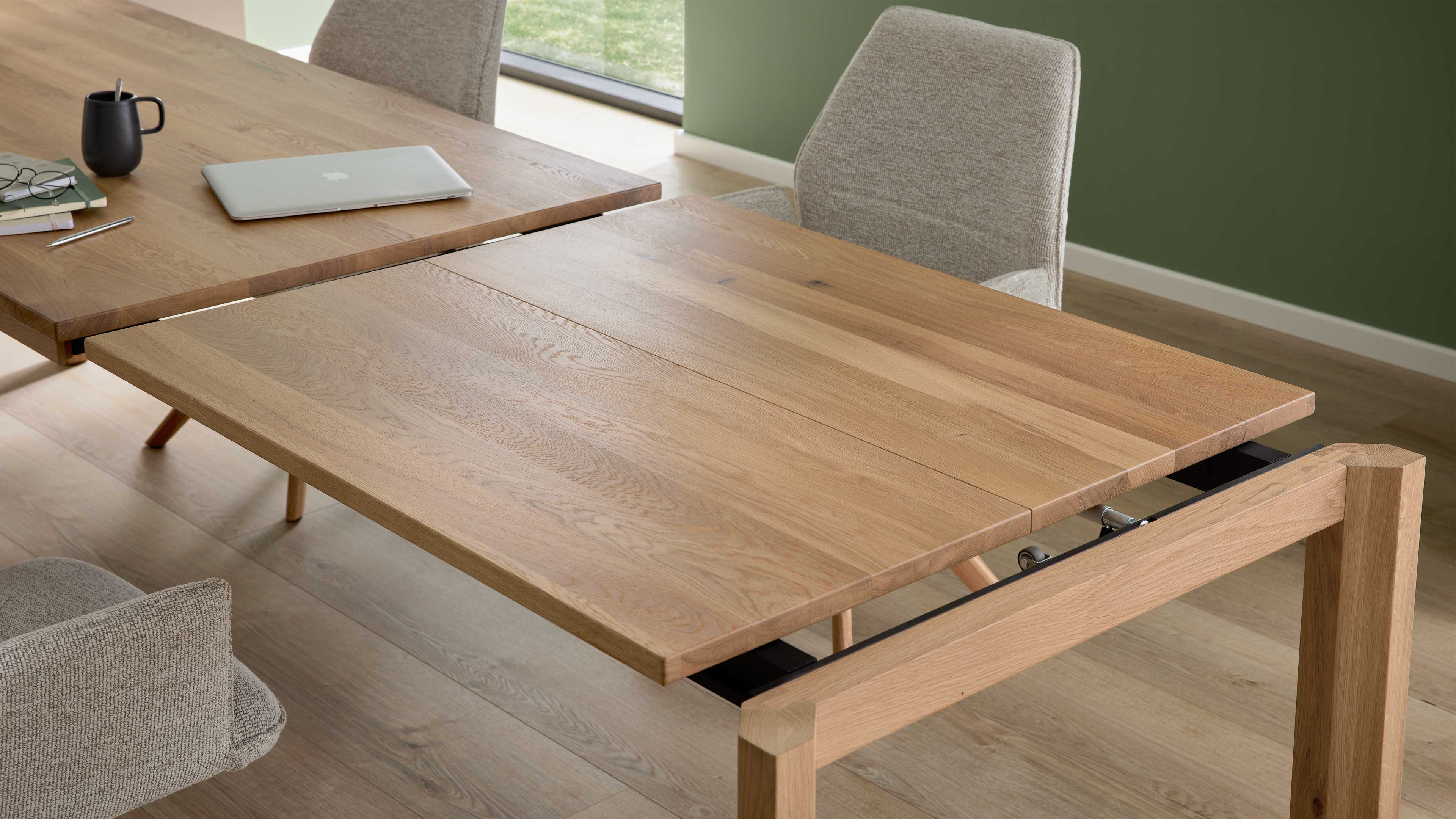 Ausziehbarer Esstisch aus hellem Holz mit ausgezogenem Verlängerungsblatt, zwei graue Polsterstühle und Laptop, Tasse.