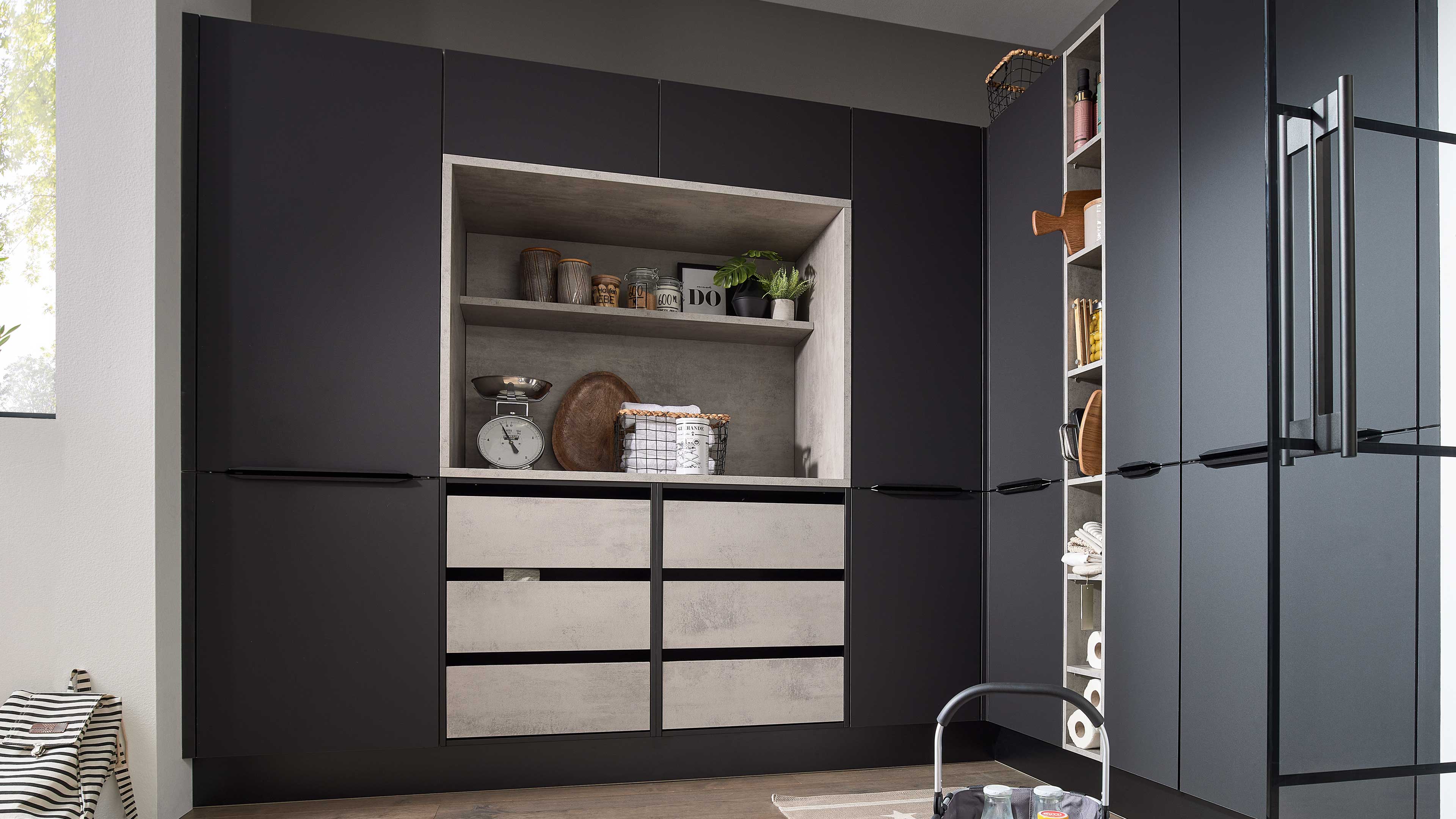 Anthrazit-Küchenfront mit zentraler hellgrauer Nische, Regalen mit Dosen und Pflanzen sowie sechs betonfarbigen Schubladen.