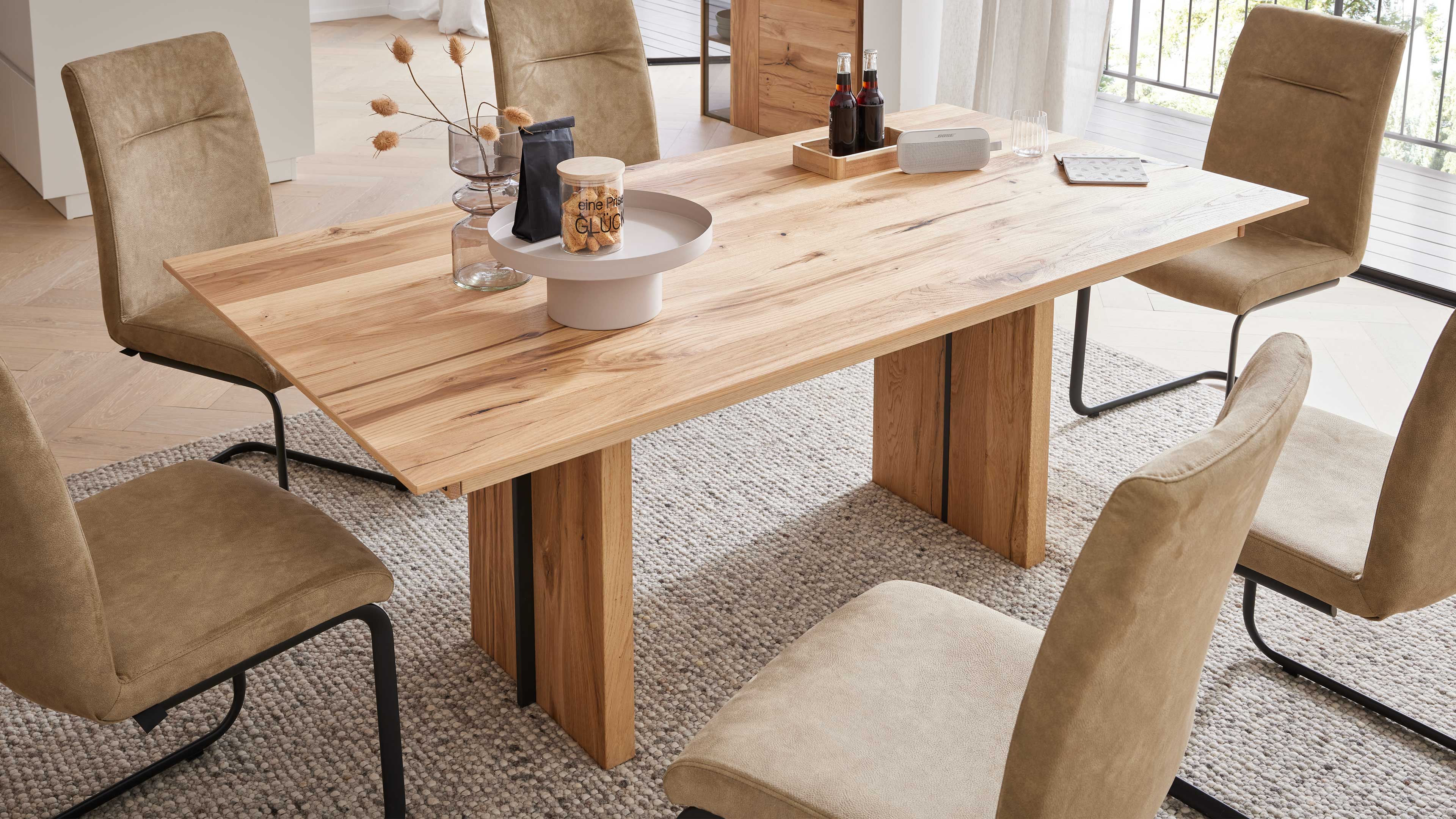 Hölzerner Tisch mit breiter Platte und zwei Beinen, umgeben von beigen Stühlen mit schwarzen Kufen und Dekoration auf der Platte.