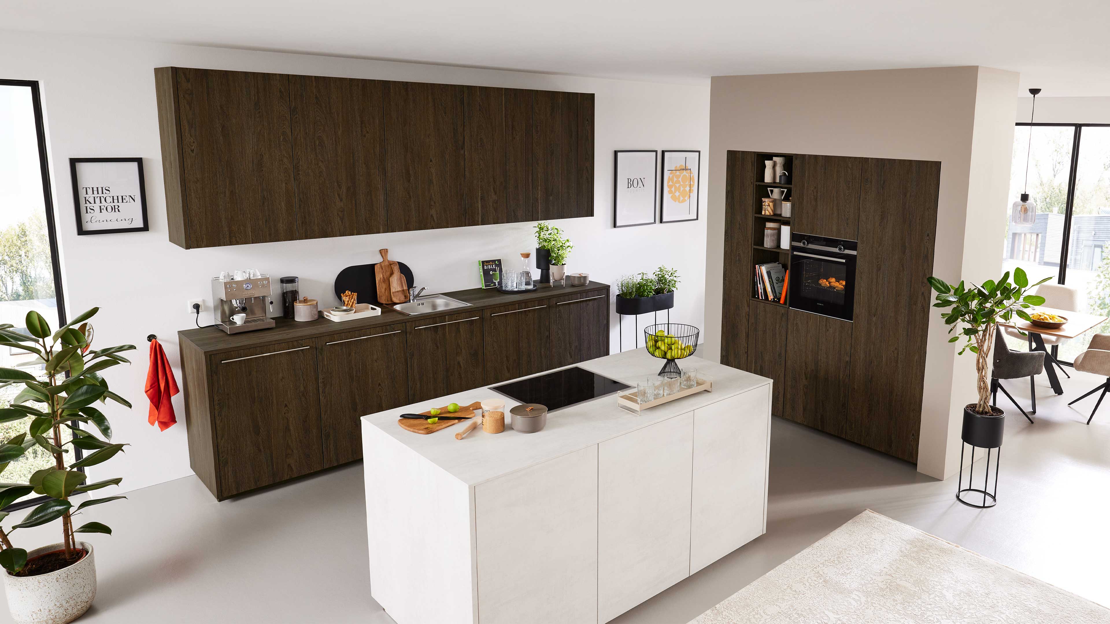 Dunkle Holz-Küchenfronten mit Aufsatzschränken und Spüle, davor eine weiße Kücheninsel mit Kochfeld, Obstkorb und Pflanzen.
