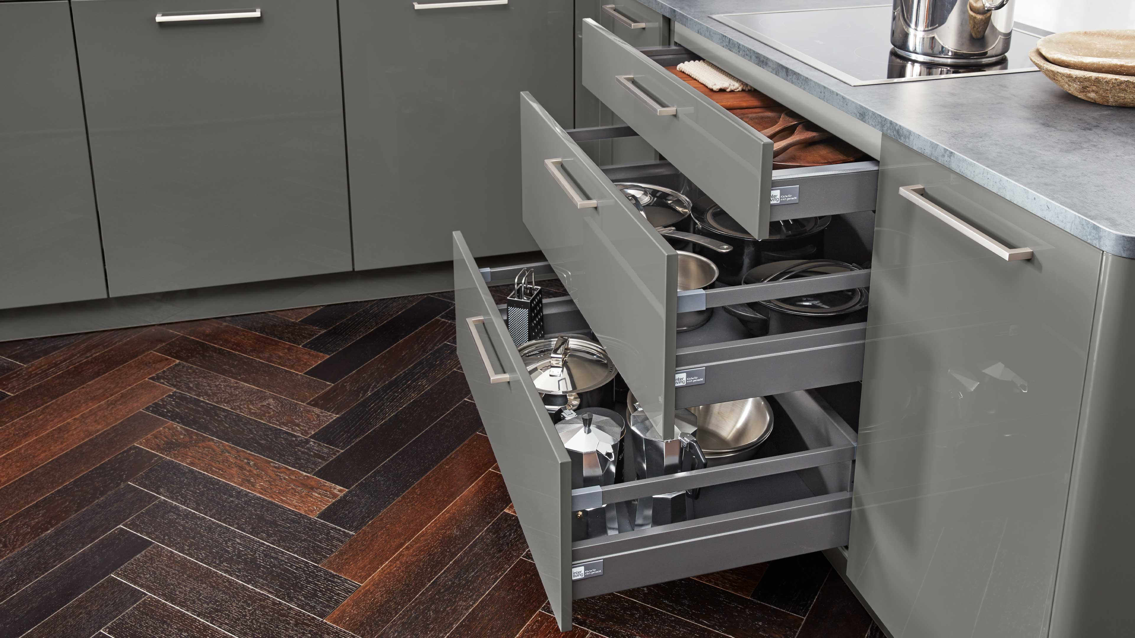 Drei geöffnete graue Küchenschubladen mit glänzenden Fronten und Metallgriffen, gefüllt mit Töpfen, Pfannen und Küchenutensilien.