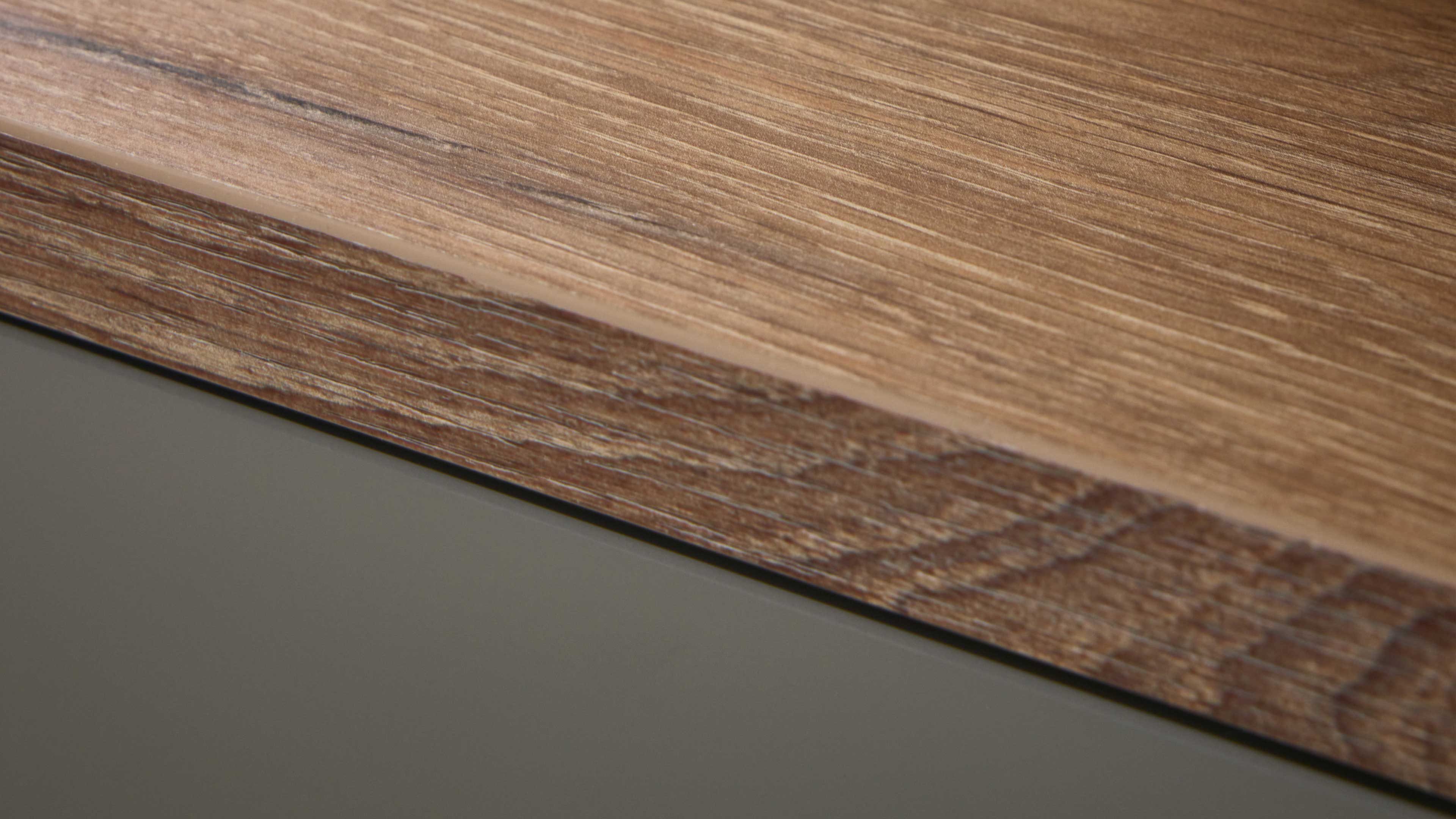 Nahaufnahme einer Holzplatte mit hellbrauner, längs verlaufender Maserung, dunklerer Kante und darunter grauschwarzer Oberfläche.