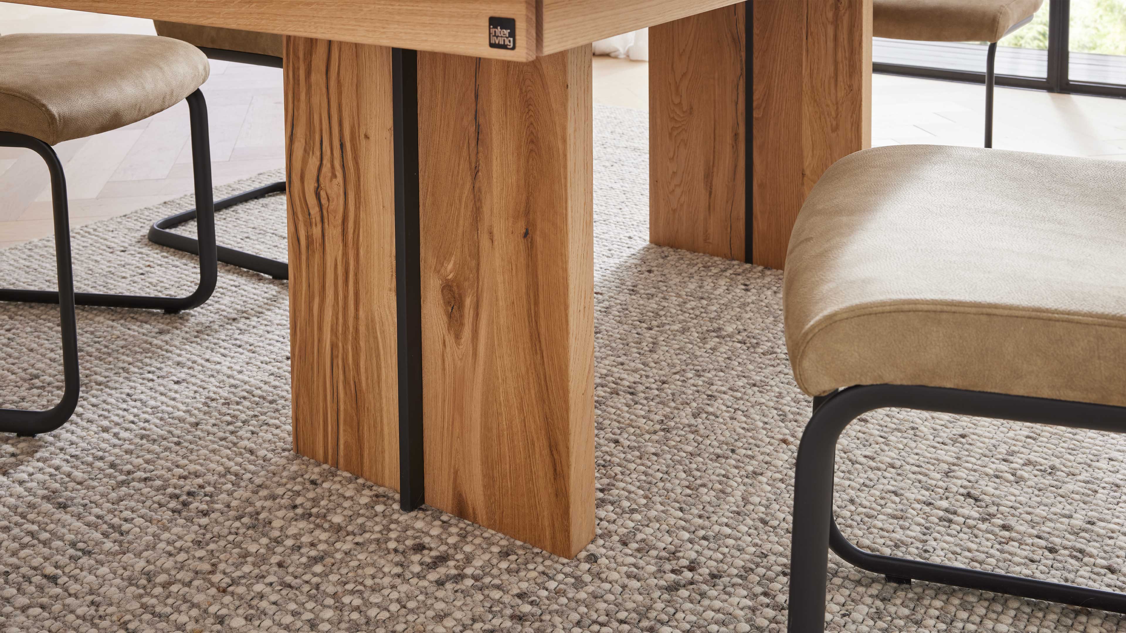 Holztischfuß mit sichtbarer Holzmaserung und schmaler schwarzer Metallblende, davor beige Polsterstühle auf grauem Teppich.