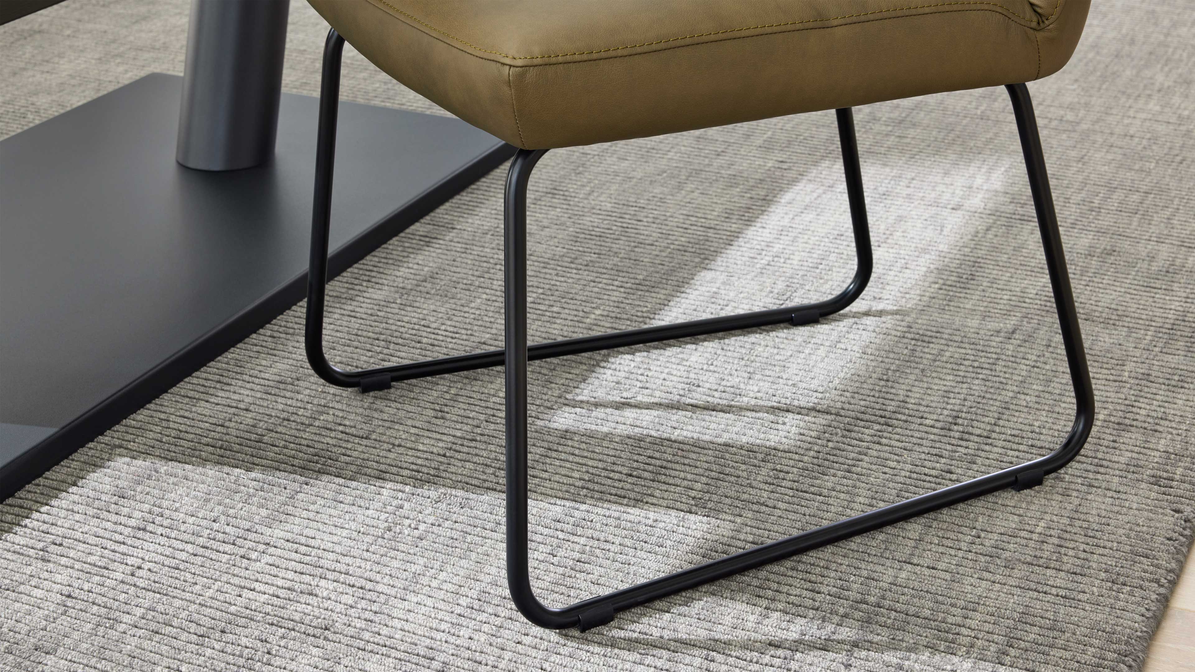 Sitzfläche in olivgrün mit sichtbaren Nähten auf schwarzem Metall-Schlittengestell, auf hellgrauem geripptem Teppich neben Tischfuß.