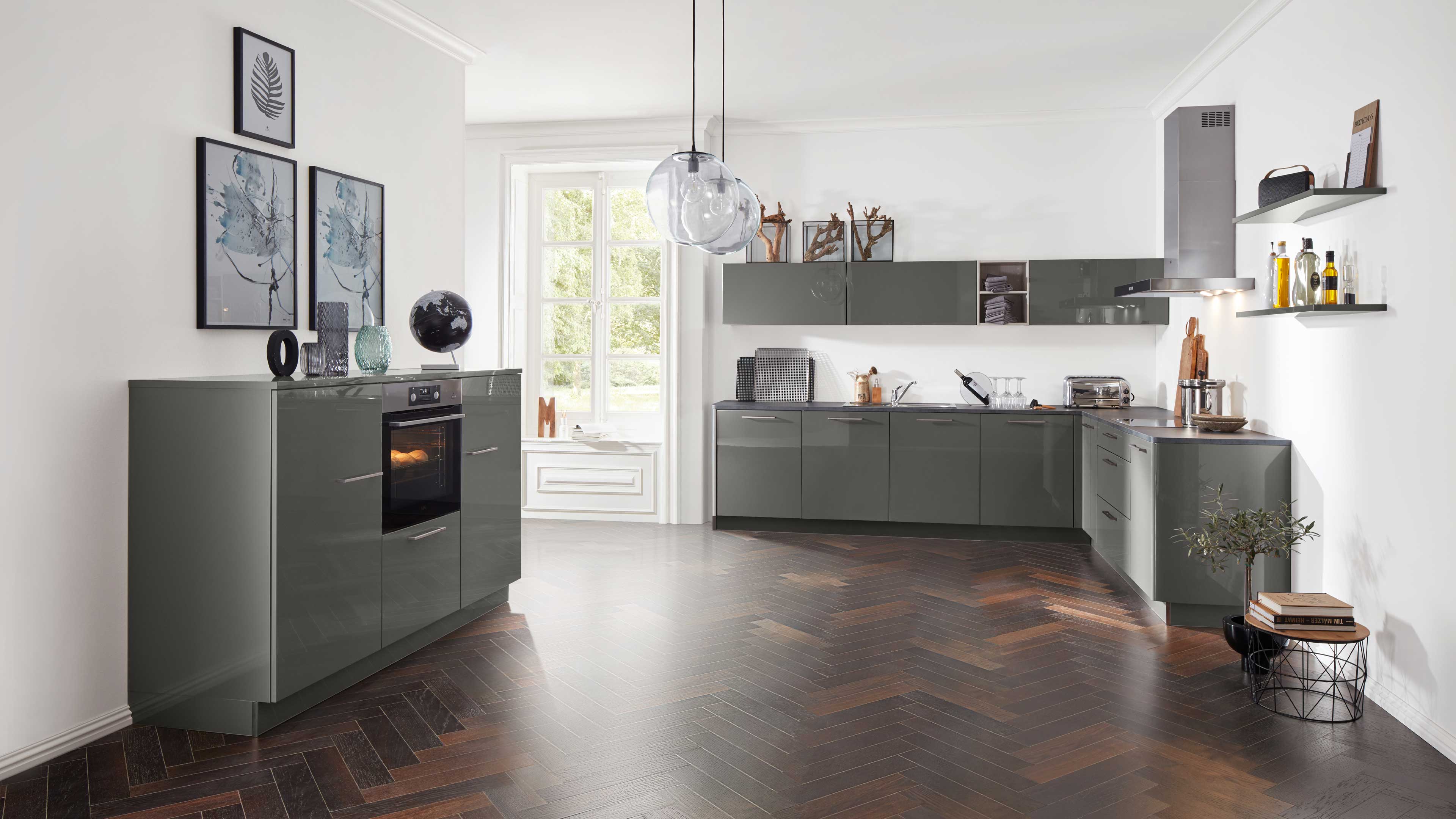 L-förmige Küchenzeile mit glänzenden graugrünen Fronten, integriertem Ofen, wandhängenden Oberschränken und dunklem Fischgrätboden.