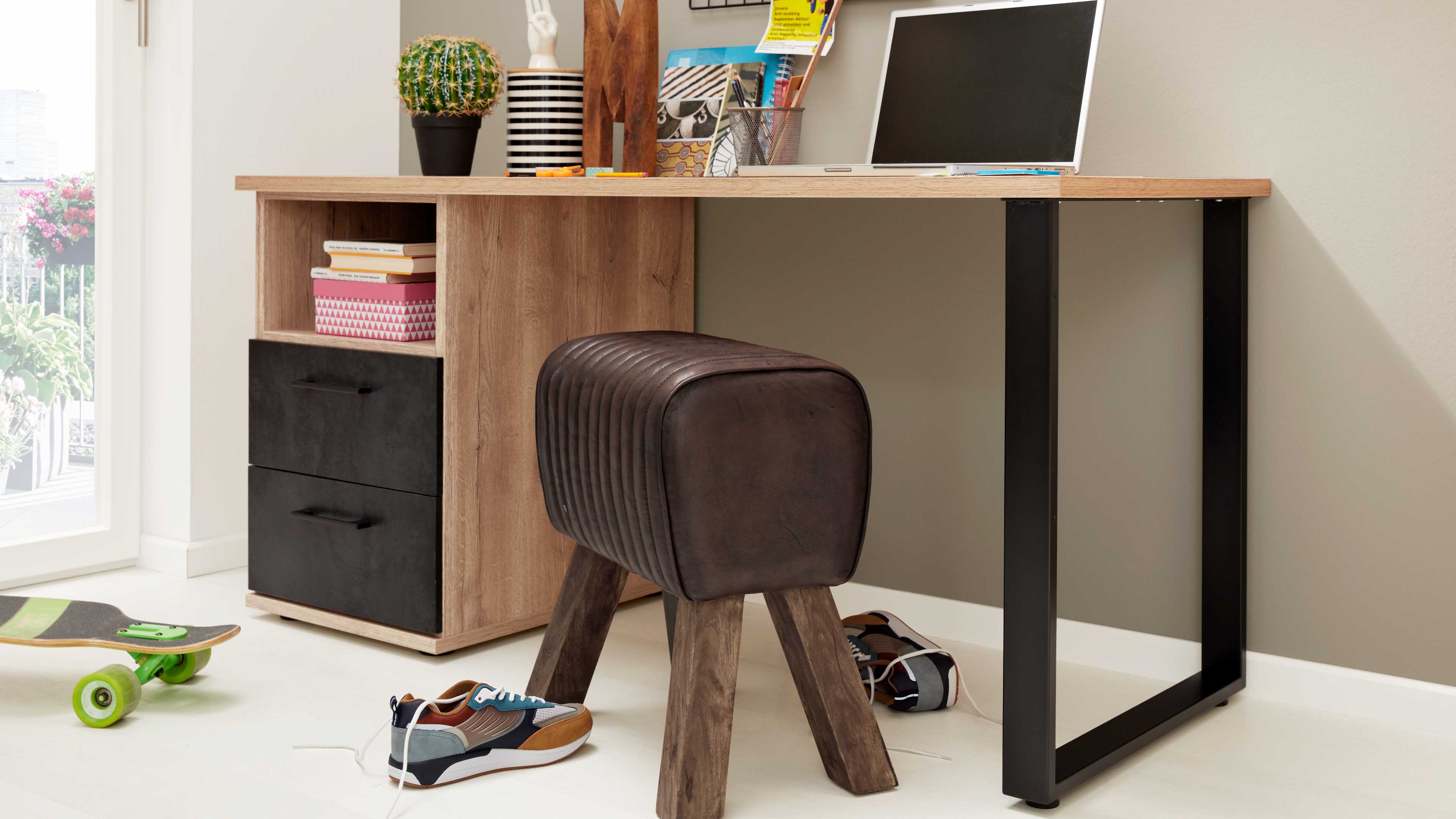 Gepolsterter, dunkelbrauner Hocker mit geripptem Leder und vier Holzbeinen vor einem Holztisch mit schwarzem Metallbein und Regalfach.