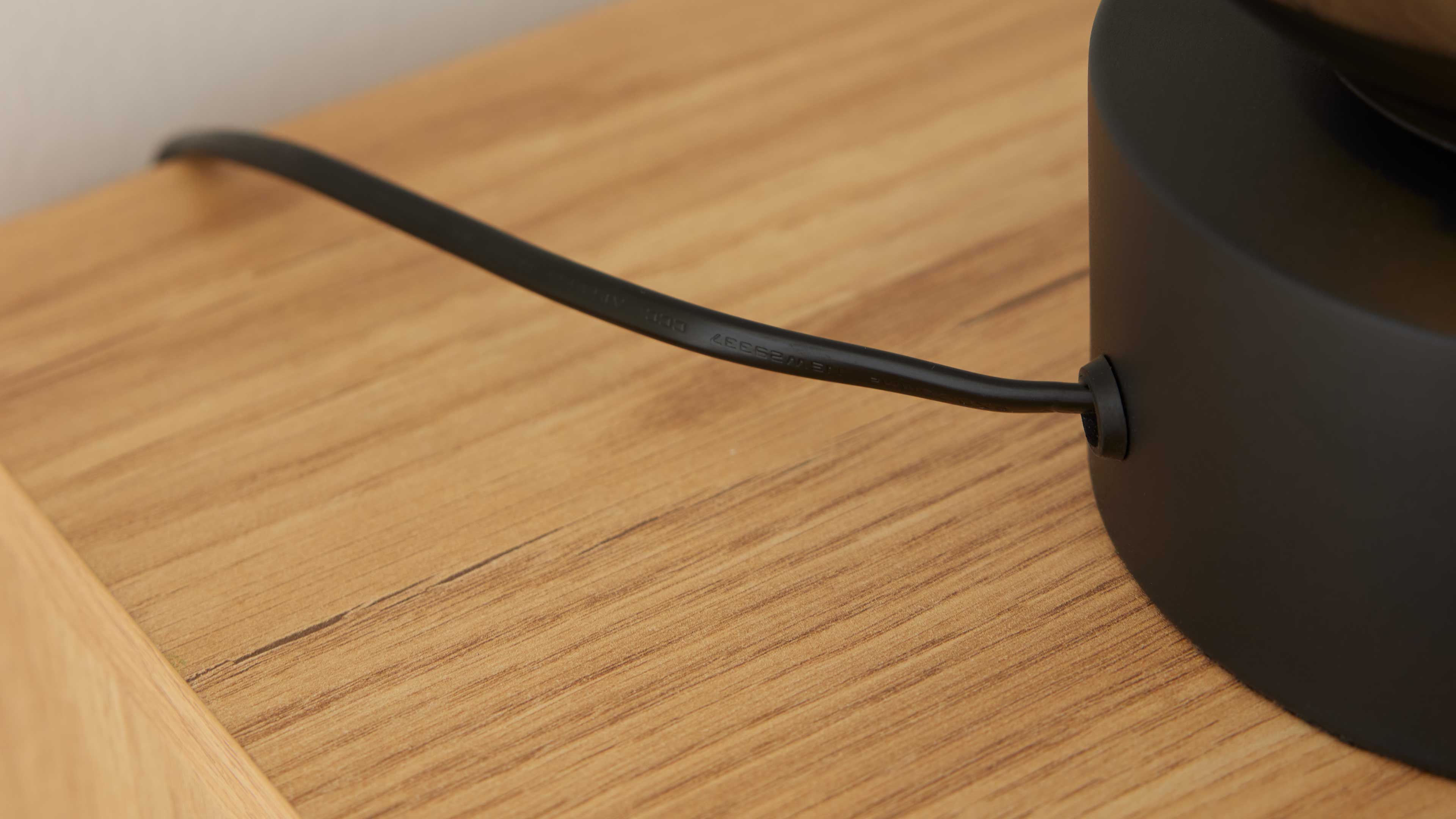 Schwarzer zylindrischer Lampenfuß mit seitlich eingeführtem schwarzem Kabel auf hellbraunem Holztisch mit sichtbarer Maserung.