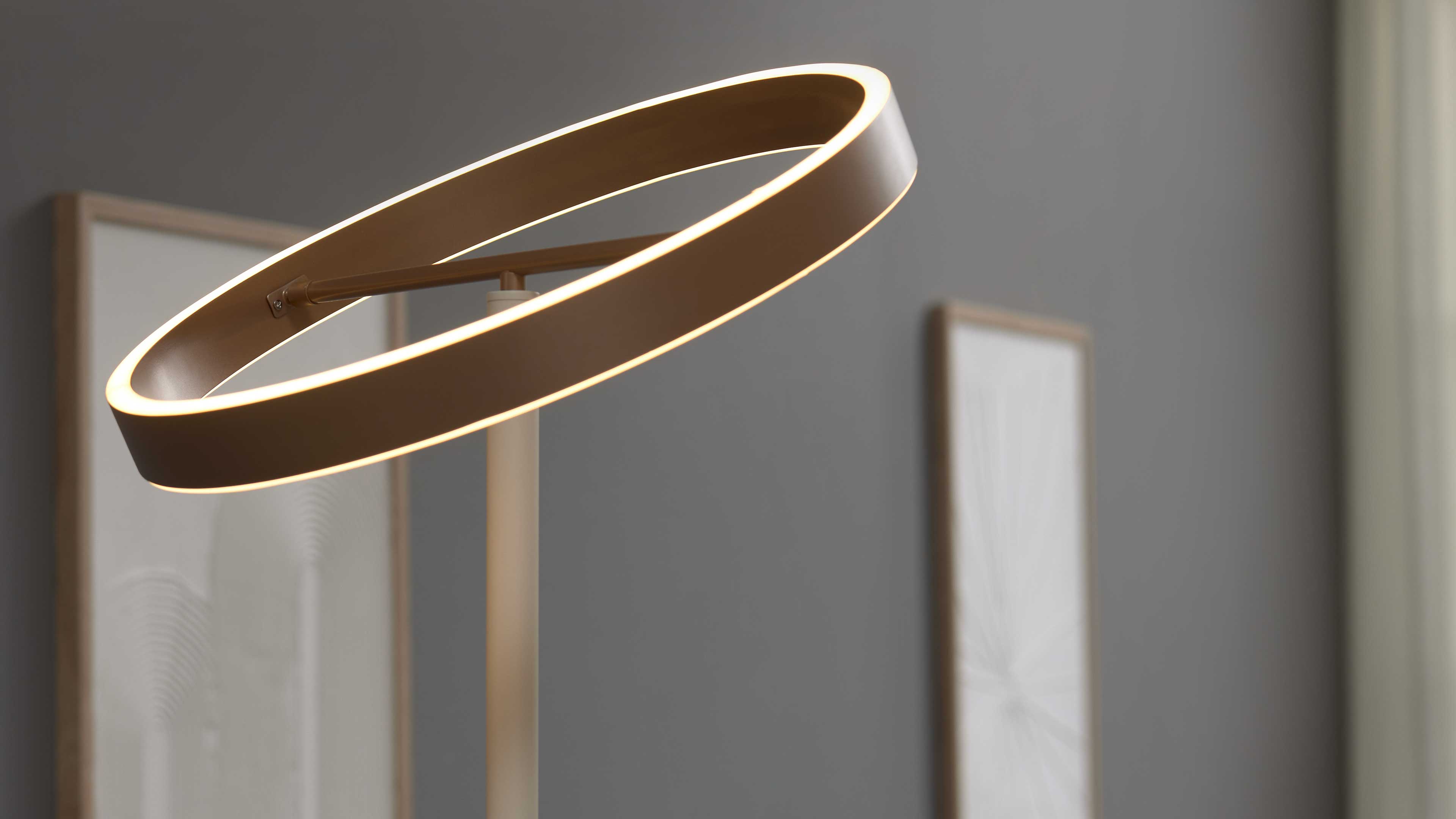 Runde, schräg gestellte Ringlampe mit leuchtendem Rand an schlankem Ständer vor grauer Wand und zwei unscharfen Bilderrahmen.