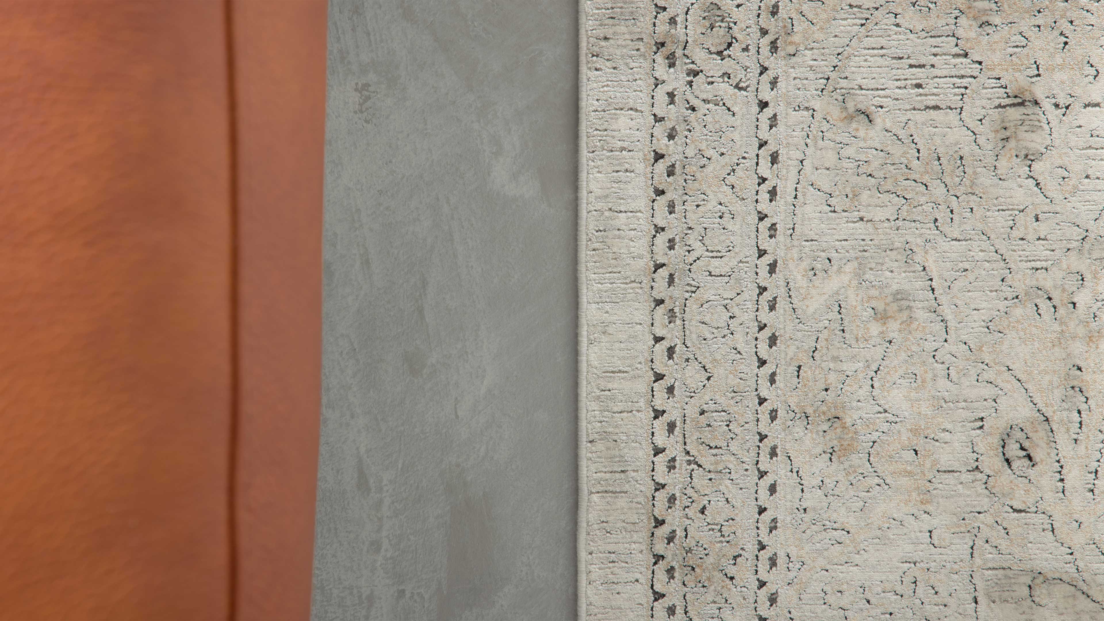 Hellgemusterter Teppich mit dekorativer Bordüre rechts, daneben glatter grauer Betonboden und links braunes Lederpolster.