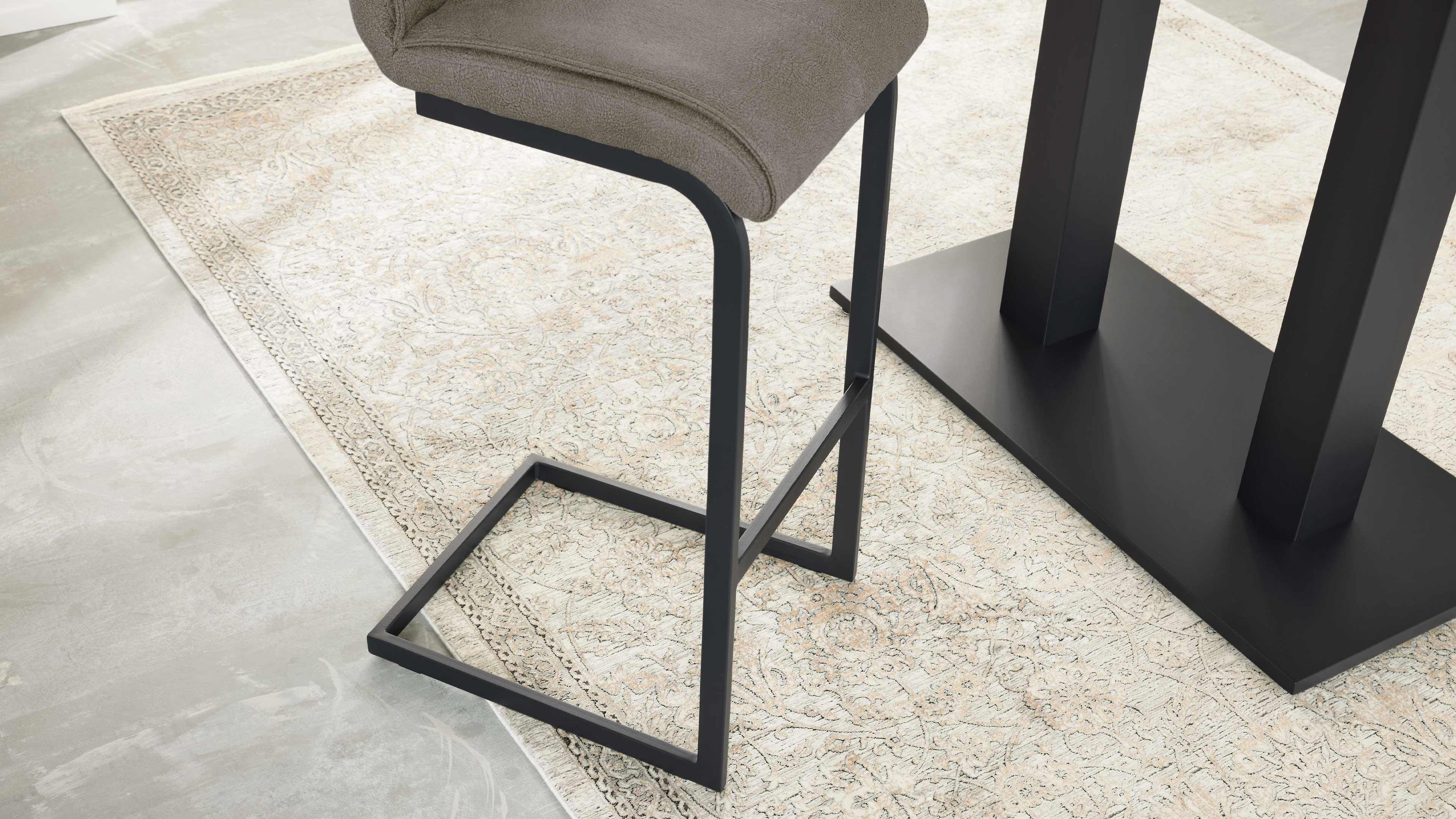 Sitzfläche mit taupefarbenem Bezug und schwarzem, kantigem Metallgestell neben schwarzem Tischfuß auf hell gemustertem Teppich.