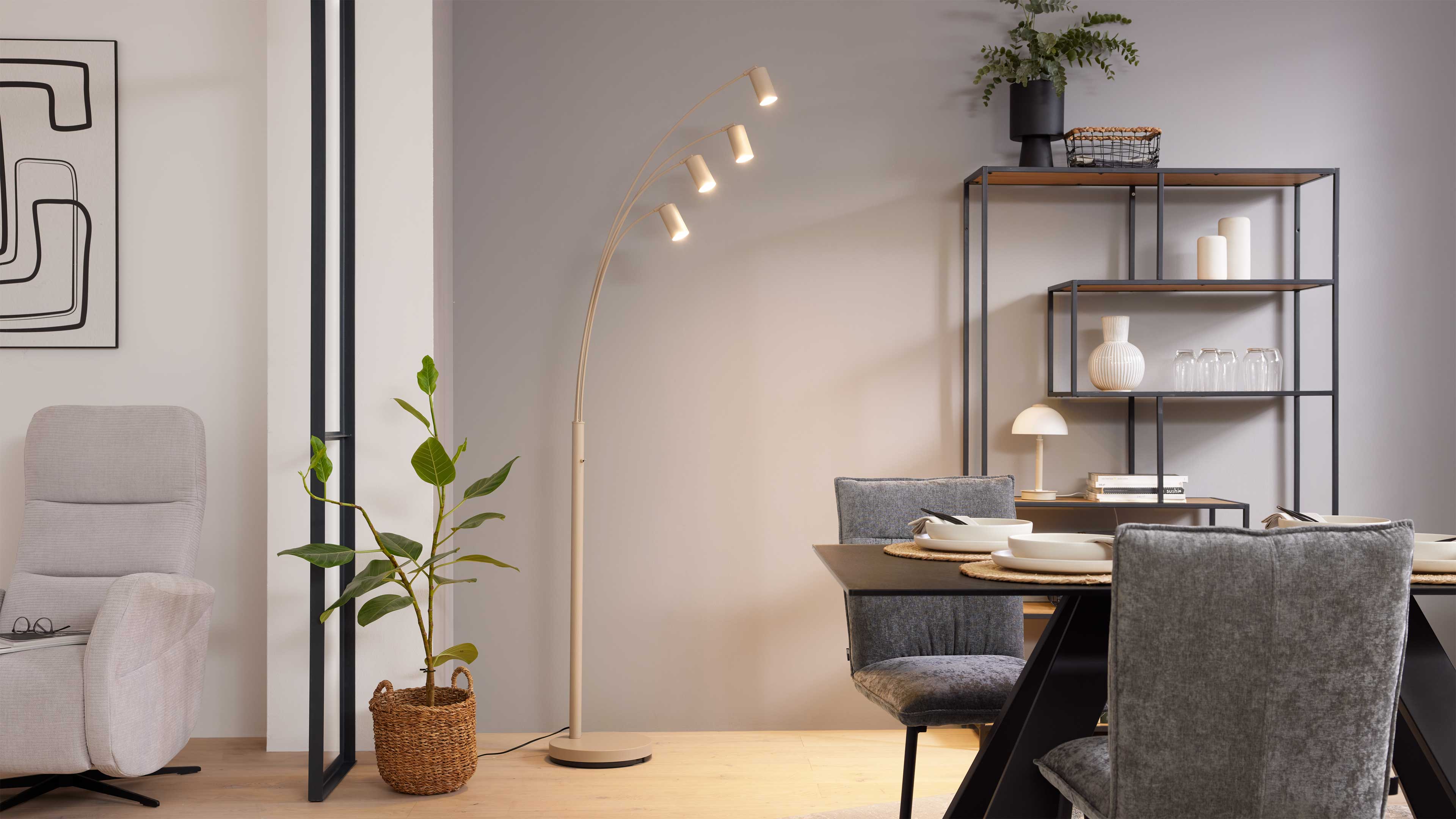 Stehende Bogenlampe mit vier leuchtenden Spots vor einer Wand, daneben Regal, Esstisch mit grauen Stühlen.