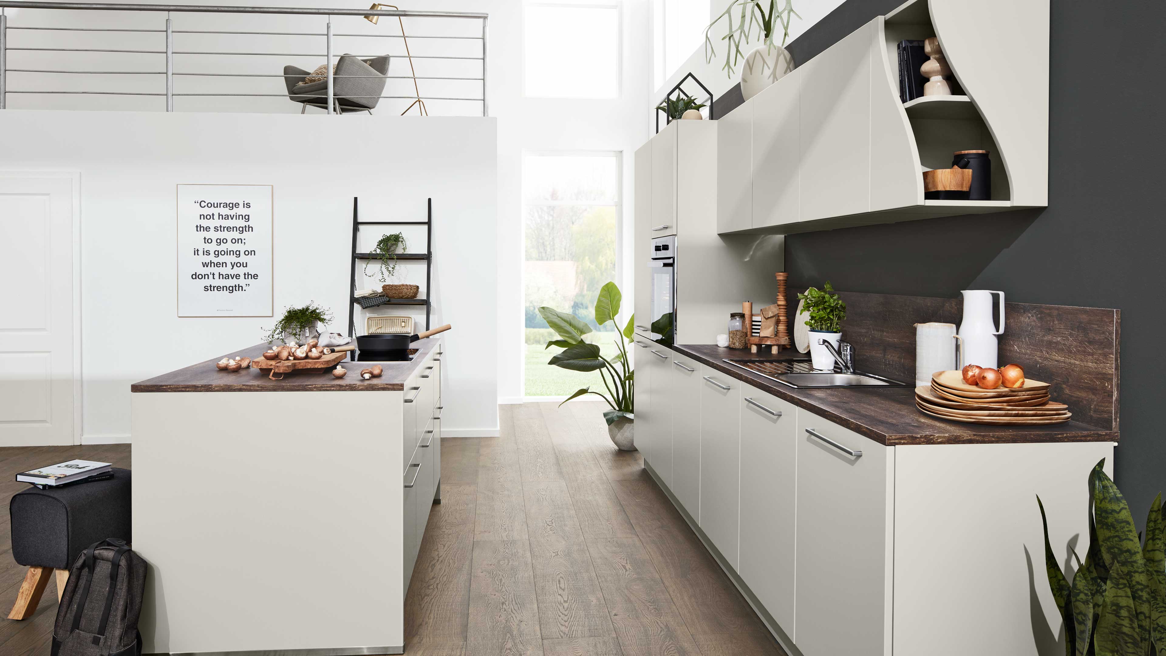 Helle Kücheninsel mit dunkler Holzarbeitsplatte und Zwiebeln, rechts lange Küchenzeile mit Spüle, Ofen und Pflanzen.