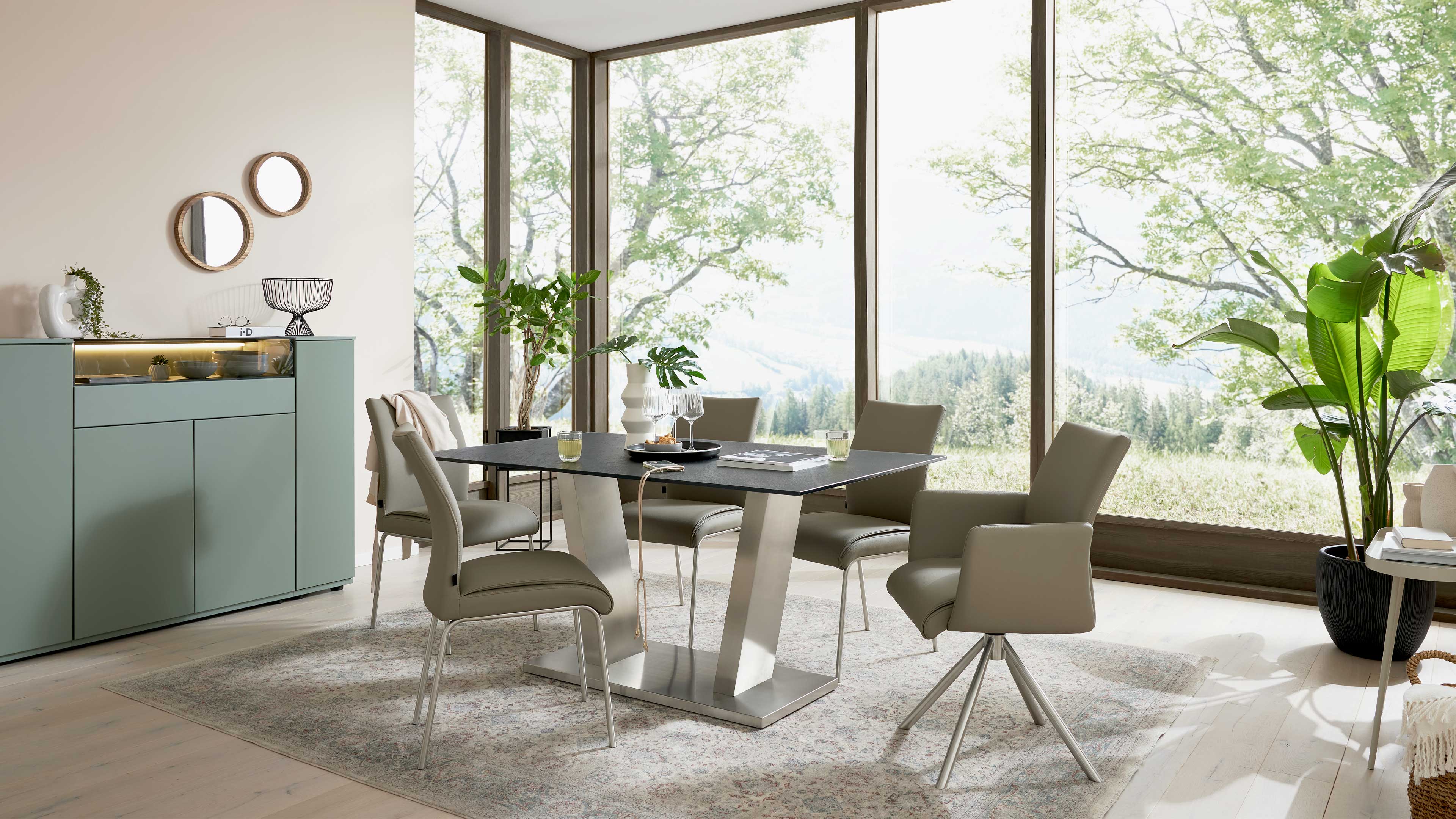 Rechteckiger Esstisch mit schwarzer Platte auf Metallgestell, umgeben von graubraunen Stühlen, links Sideboard und große Fenster mit Pflanzen.