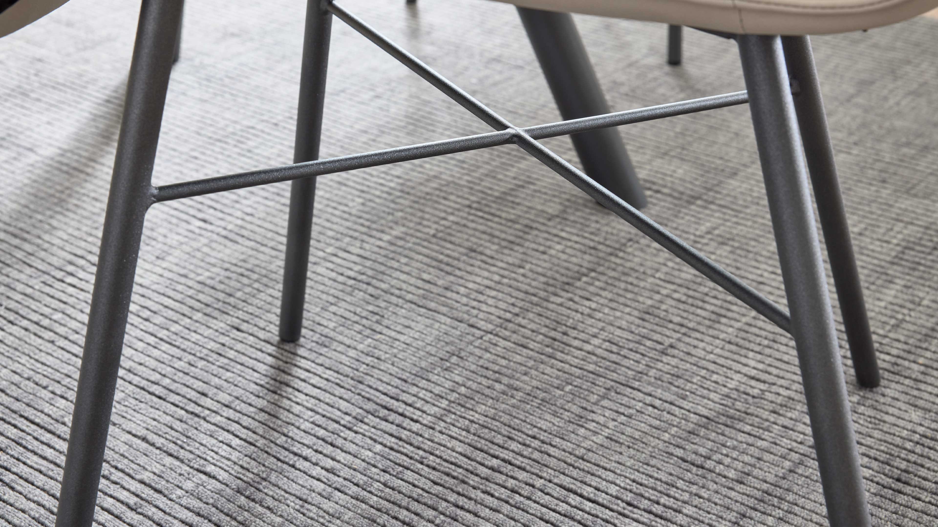Unterseite eines Stuhls mit vier schrägen, dunkelgrauen Metallbeinen und gekreuzten Querstangen auf strukturiertem hellgrauen Teppich.