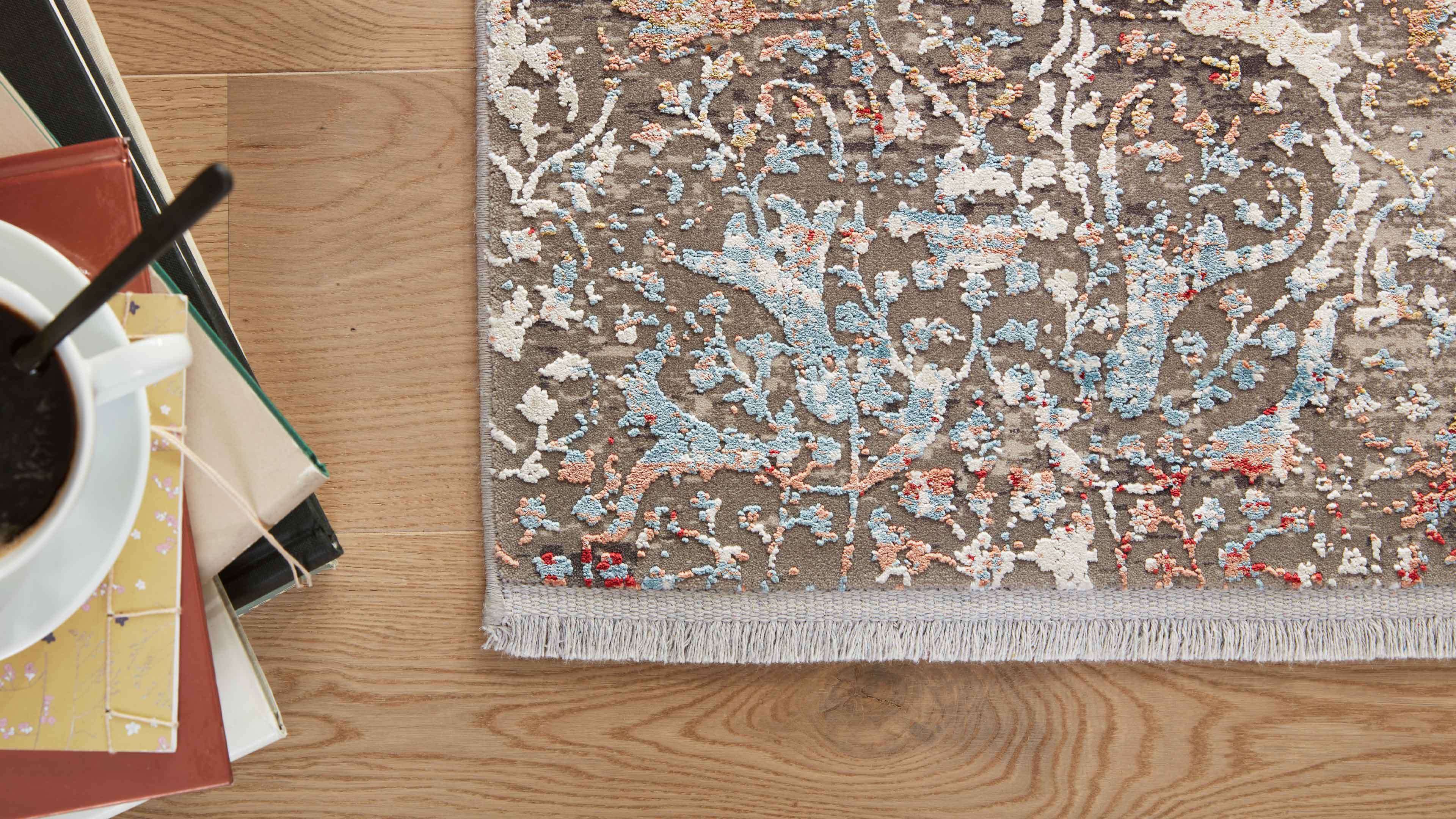 Teppich mit mehrfarbigem, floralen Reliefmuster und kurzen Fransen liegt auf hellem Holzfußboden; links Bücherstapel und Kaffeetasse.