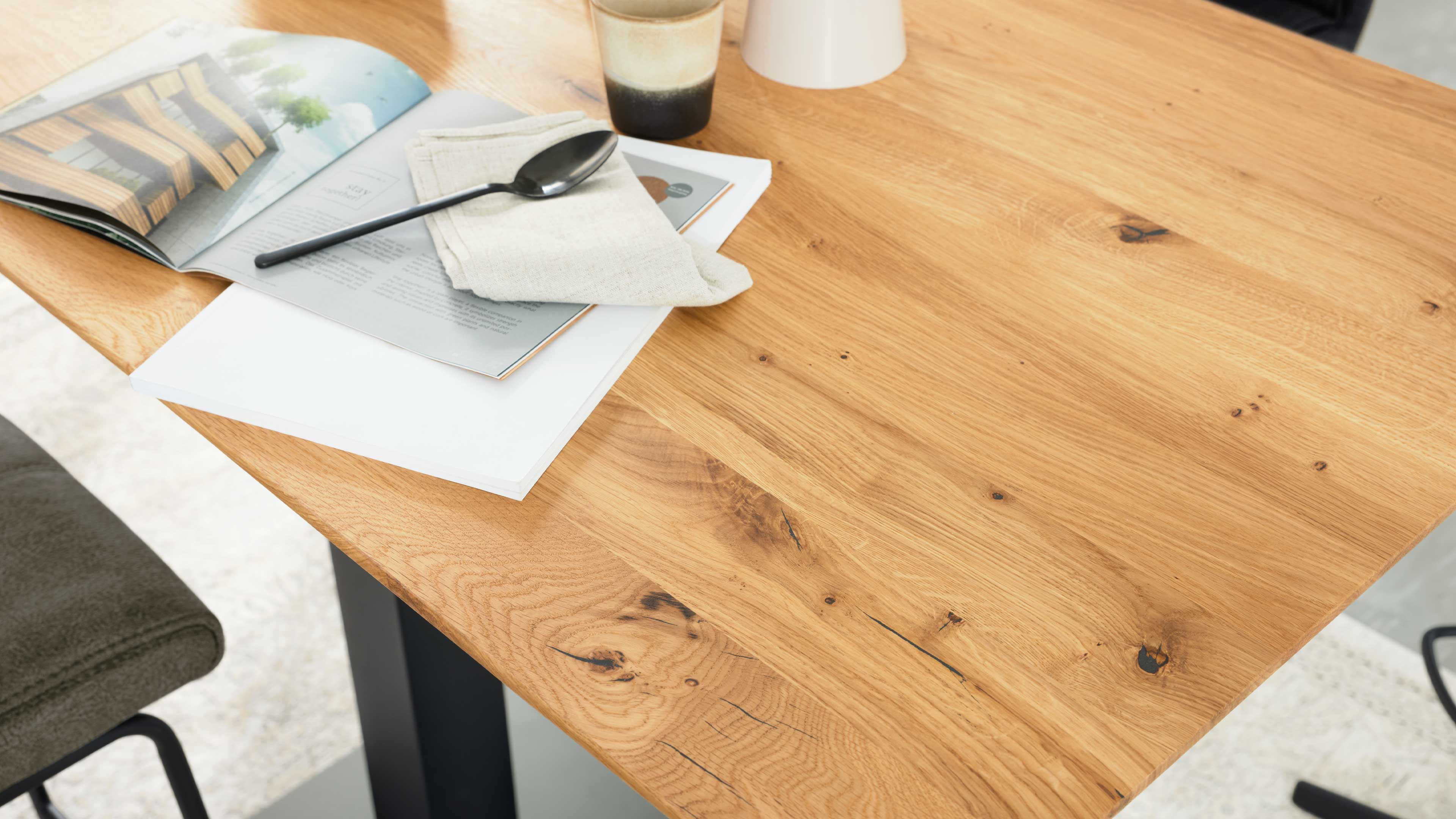 Holztischplatte mit sichtbarer Maserung und Astlöchern, darauf Magazin, gefaltete Serviette mit schwarzem Löffel und Becher.