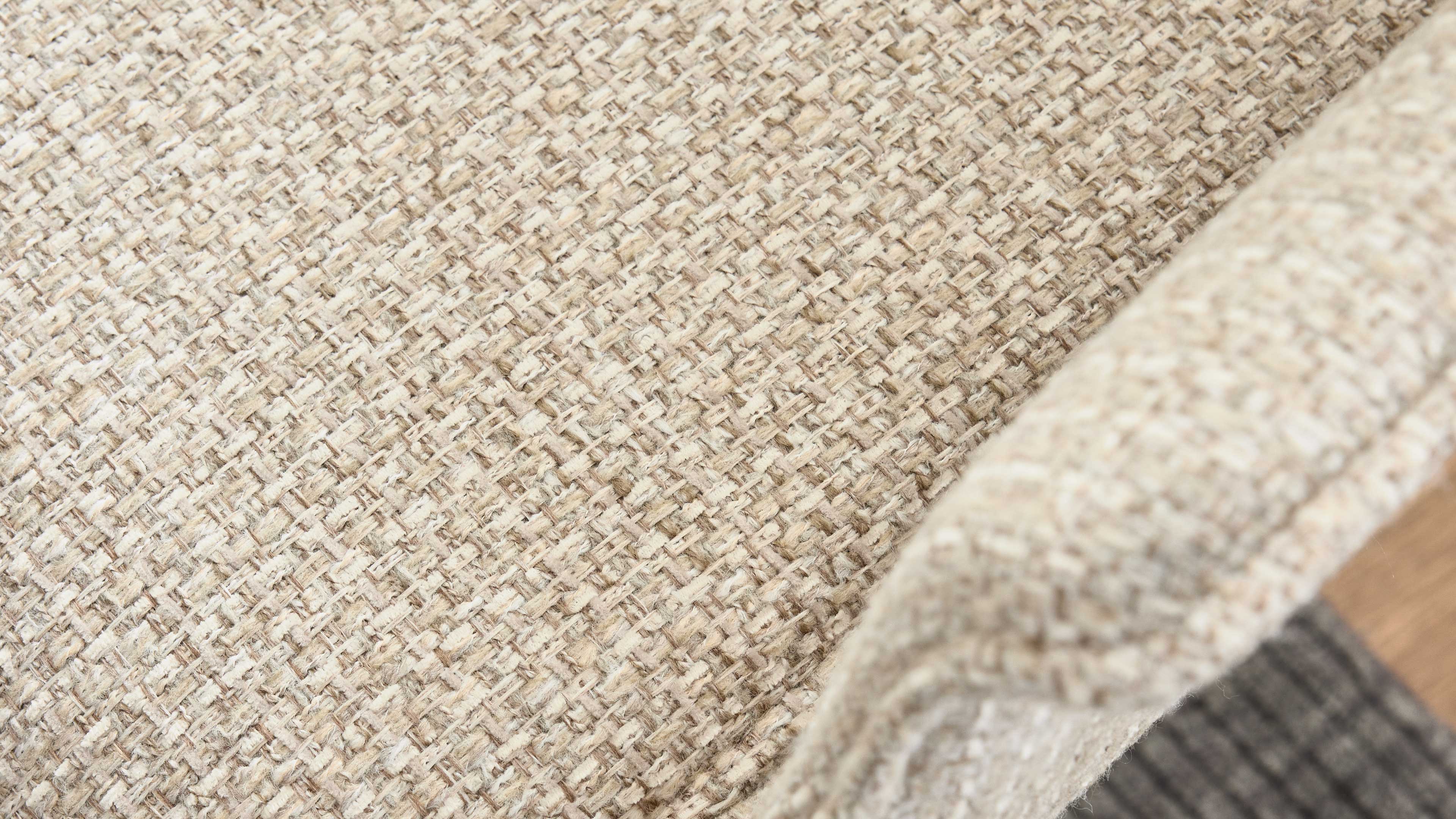 Nahaufnahme eines hellbeigen, grob gewebten Polsterstoffs auf einer abgerundeten Armlehne, rechts Bodenbelag sichtbar.