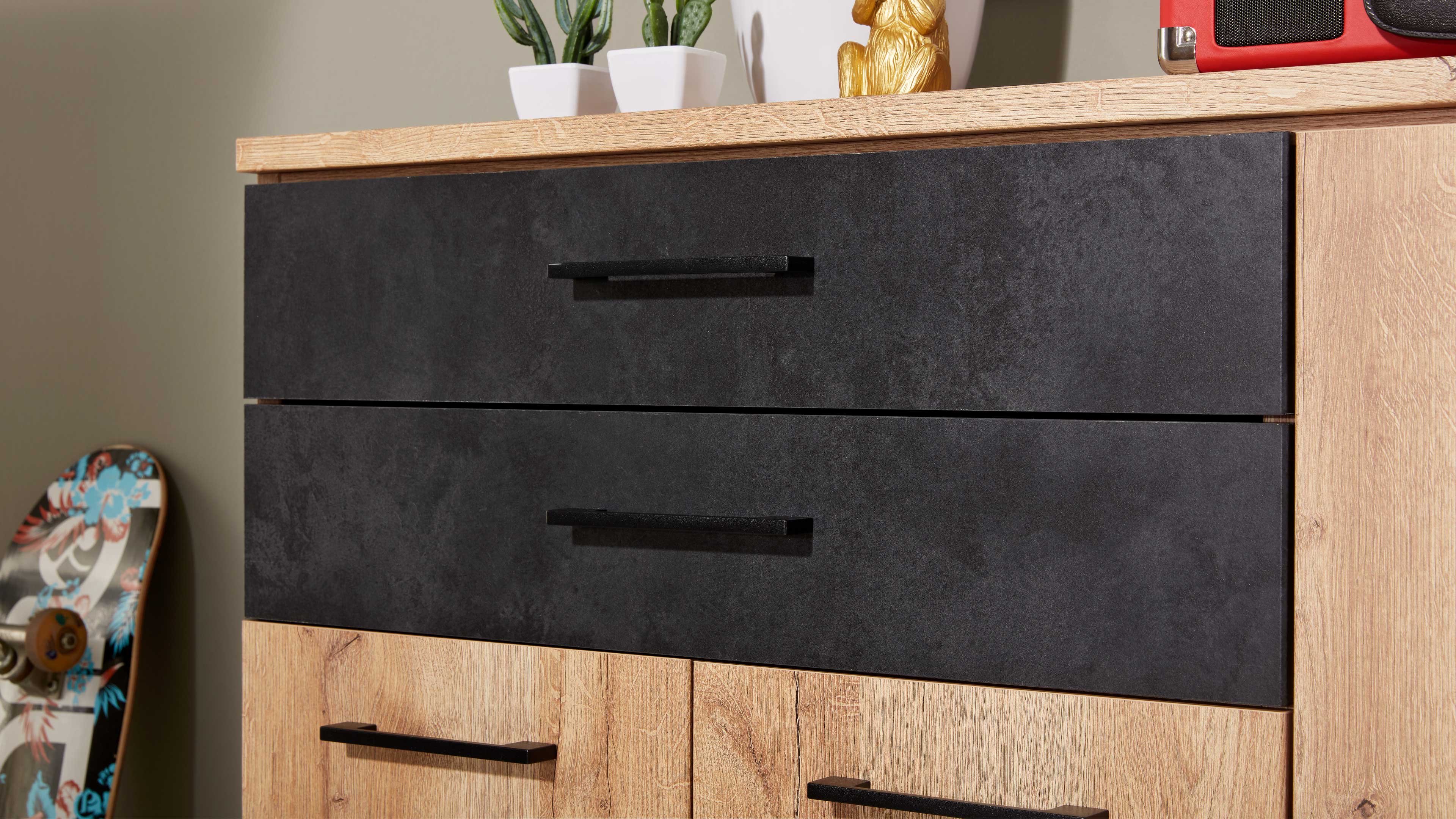 Kommode mit zwei großen dunkelgrauen Schubladen über holzfarbener Front, schwarzen Griffen, Pflanzen auf der Arbeitsplatte und Skateboard links.
