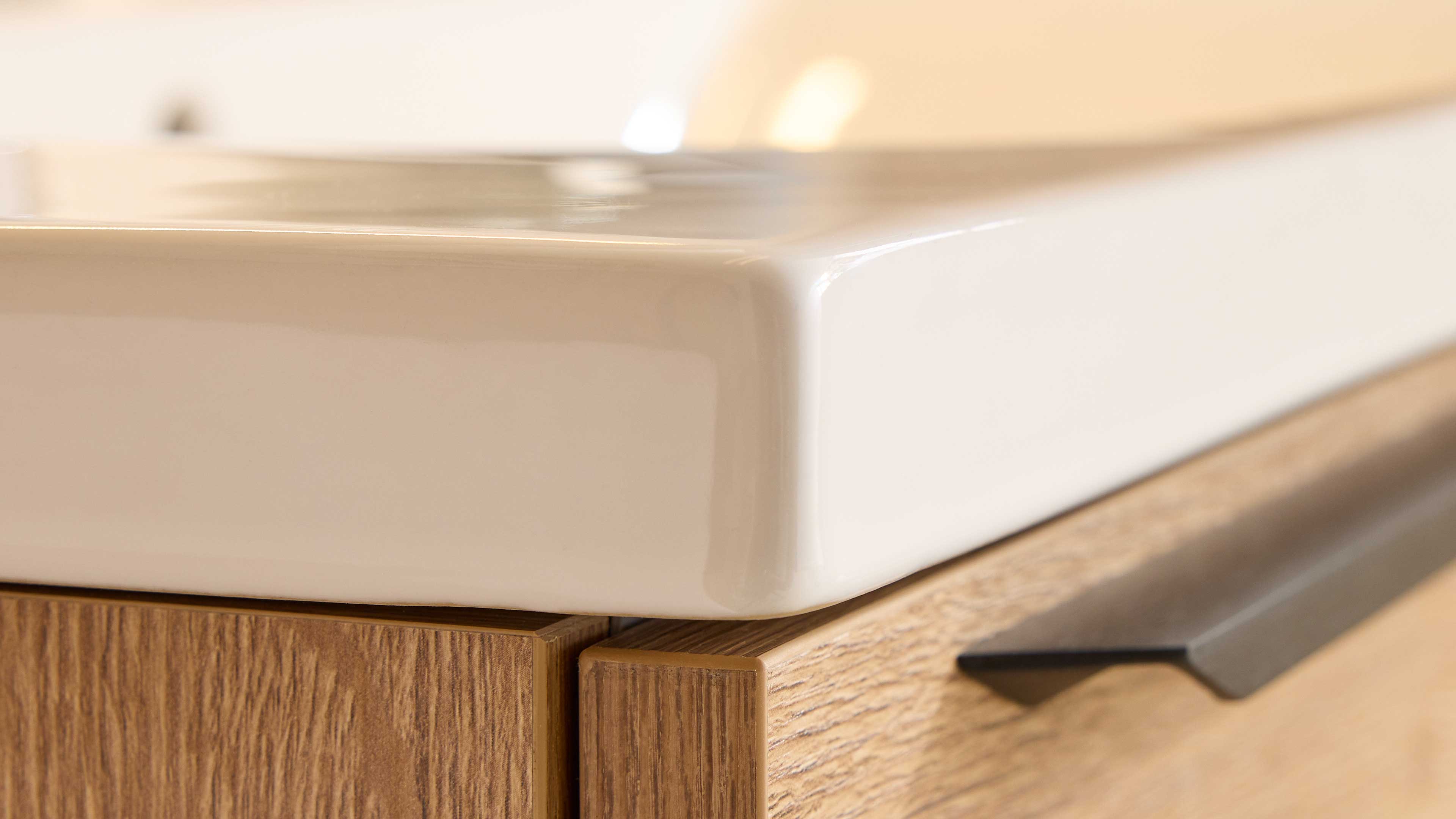 Weiße, glänzende Eckkante eines Keramikwaschbeckens, aufgesetzt auf eine Holzfront mit sichtbarer Maserung und schwarzem Griff.