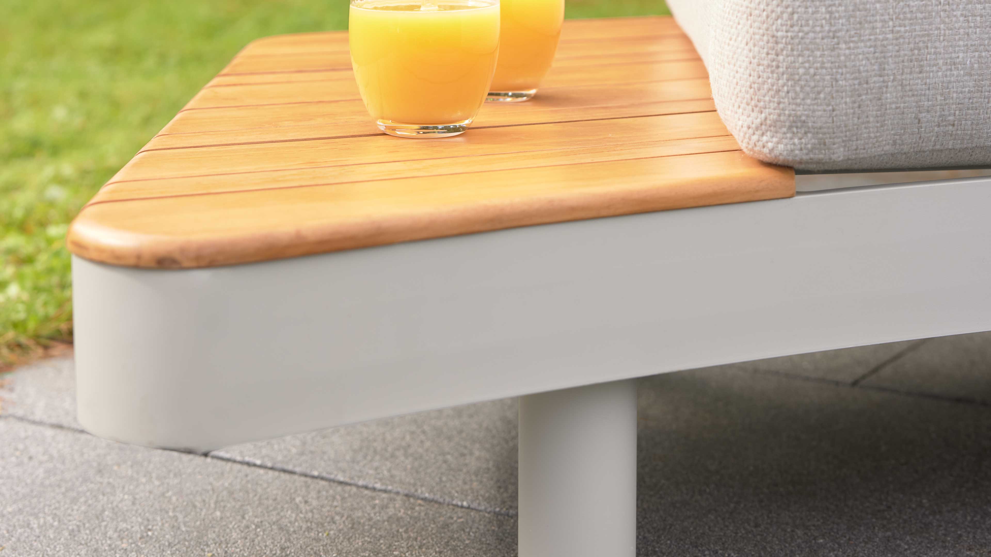 Sitzfläche mit hellen Holzlatten auf weißgrauem Gestell, beige Polsterkante und zwei Gläser Orangensaft, draußen auf Terrasse.