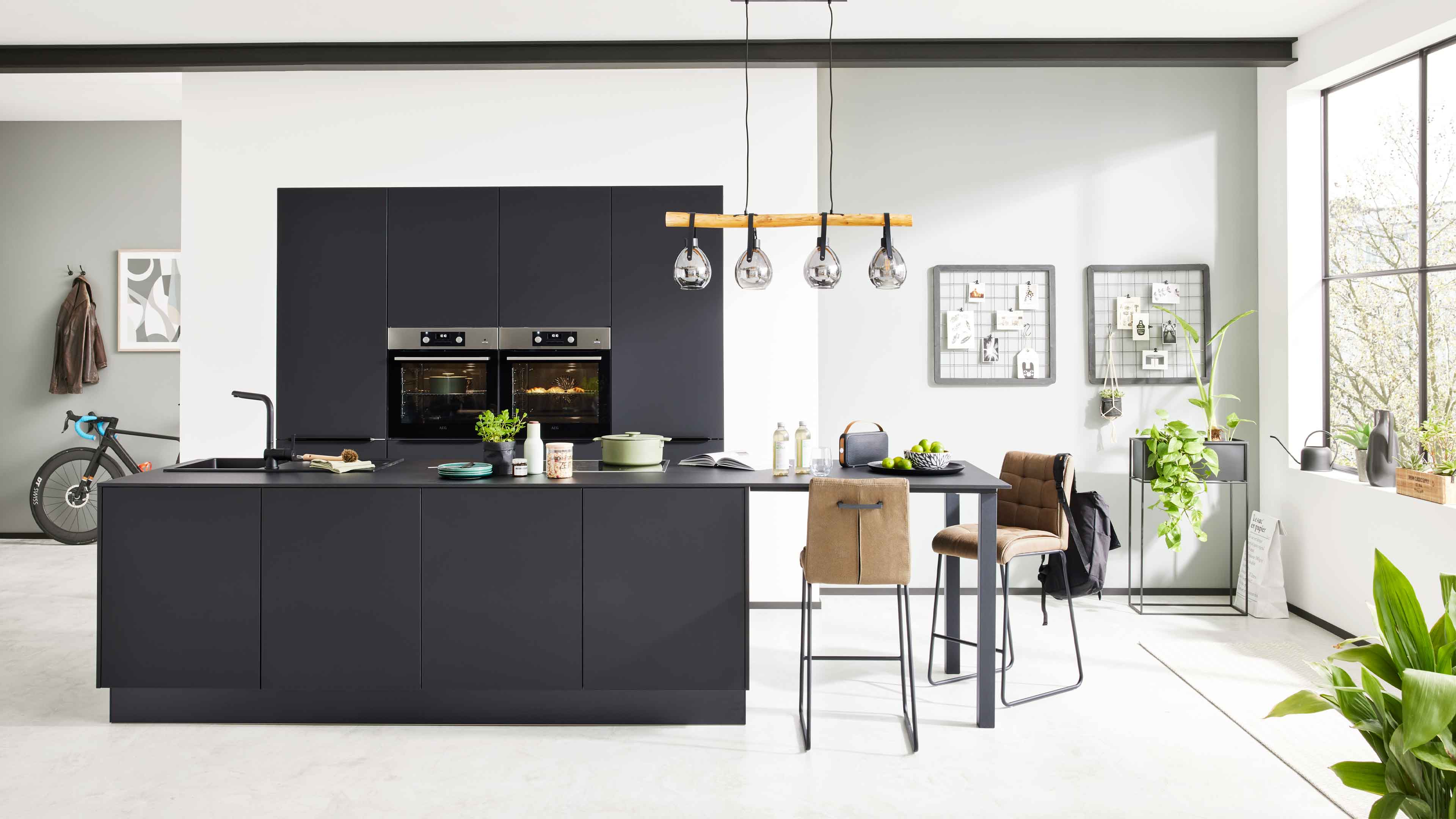 Dunkelgraue Kücheninsel mit Spüle und angeschlossenem Bartisch, zwei braune Barhocker, darüber Hängeleuchte, Pflanzen am Fenster.