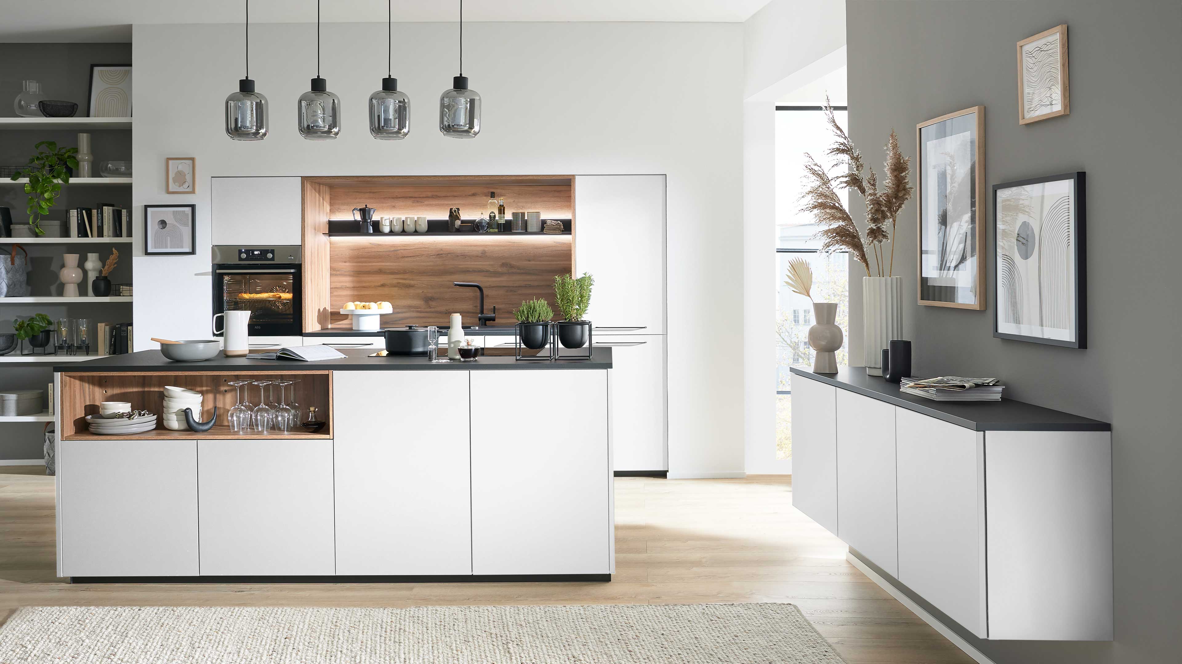 Große Kücheninsel mit weißen Fronten und offenem Holzfach mit Geschirr, vier Glashängelampen, Küchenzeile mit Ofen und Holzrückwand.