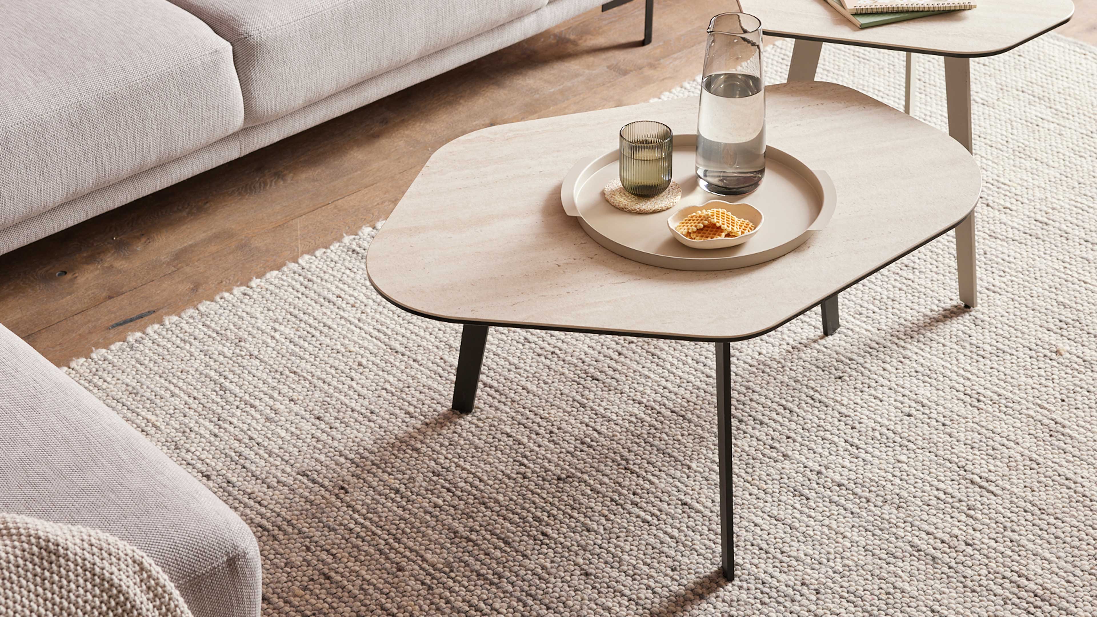 Heller, ovaler Couchtisch mit dünnen schwarzen Metallbeinen, darauf Tablett mit Karaffe, Glas und Schale; links Sofakante und Teppich.