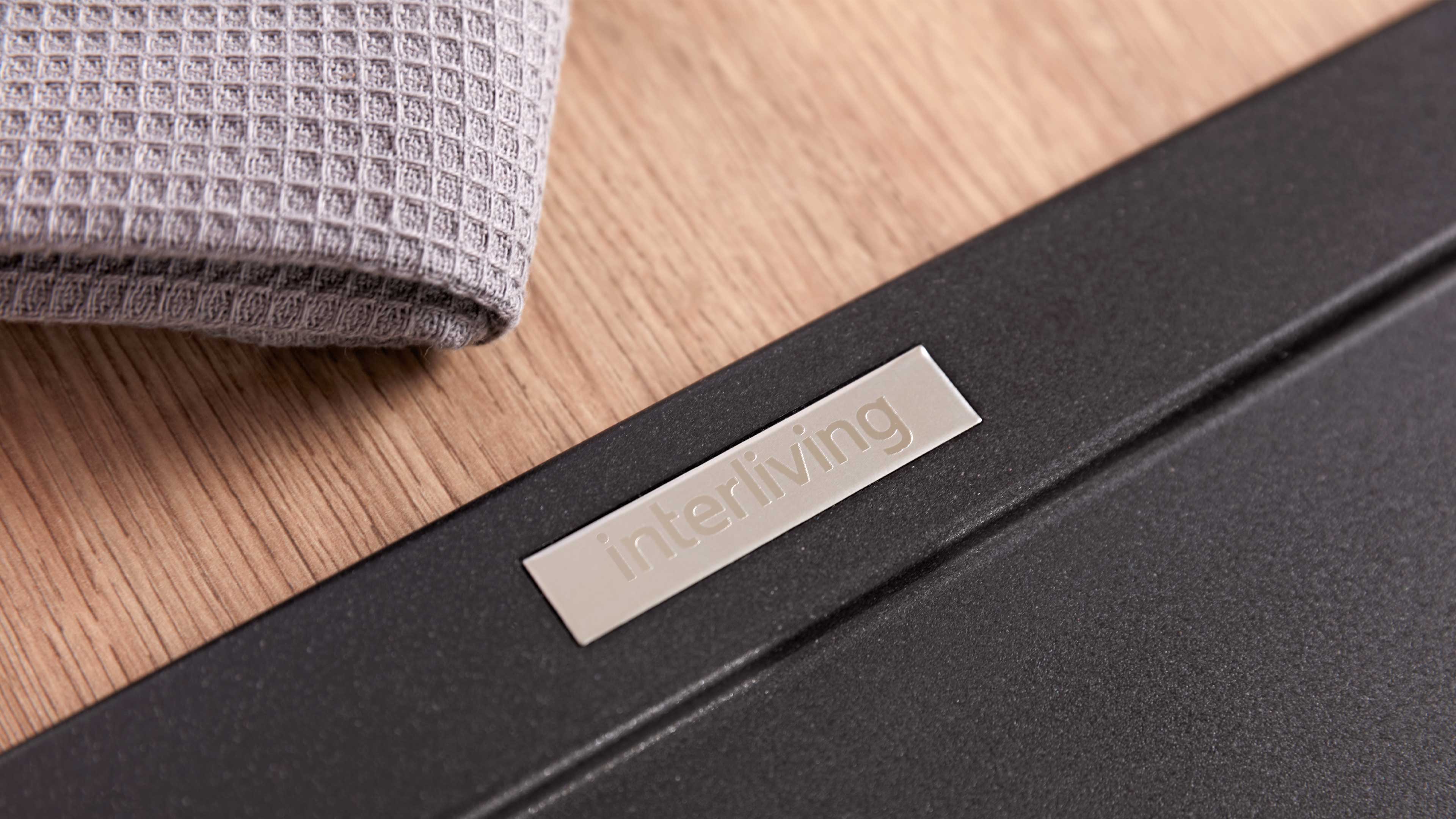 Nahaufnahme einer Eichenholzplatte mit schwarzem Metallrahmen und silberner rechteckiger Plakette mit eingeprägtem Schriftzug 'interliving'.