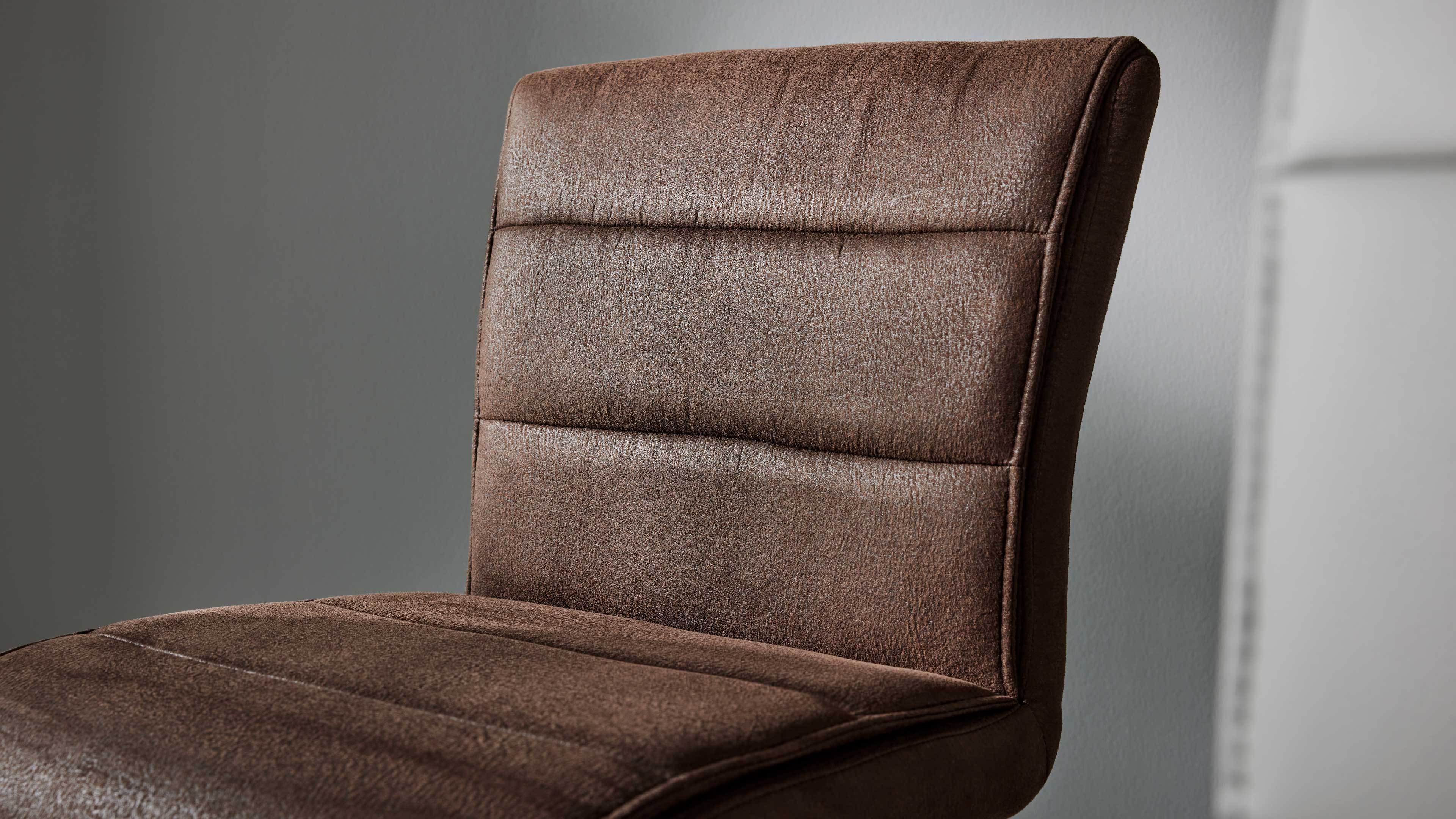 Brauner gepolsterter Stuhl in Nahaufnahme, sichtbare horizontale Steppnähte und strukturierter Bezugsstoff vor grauer Wand.