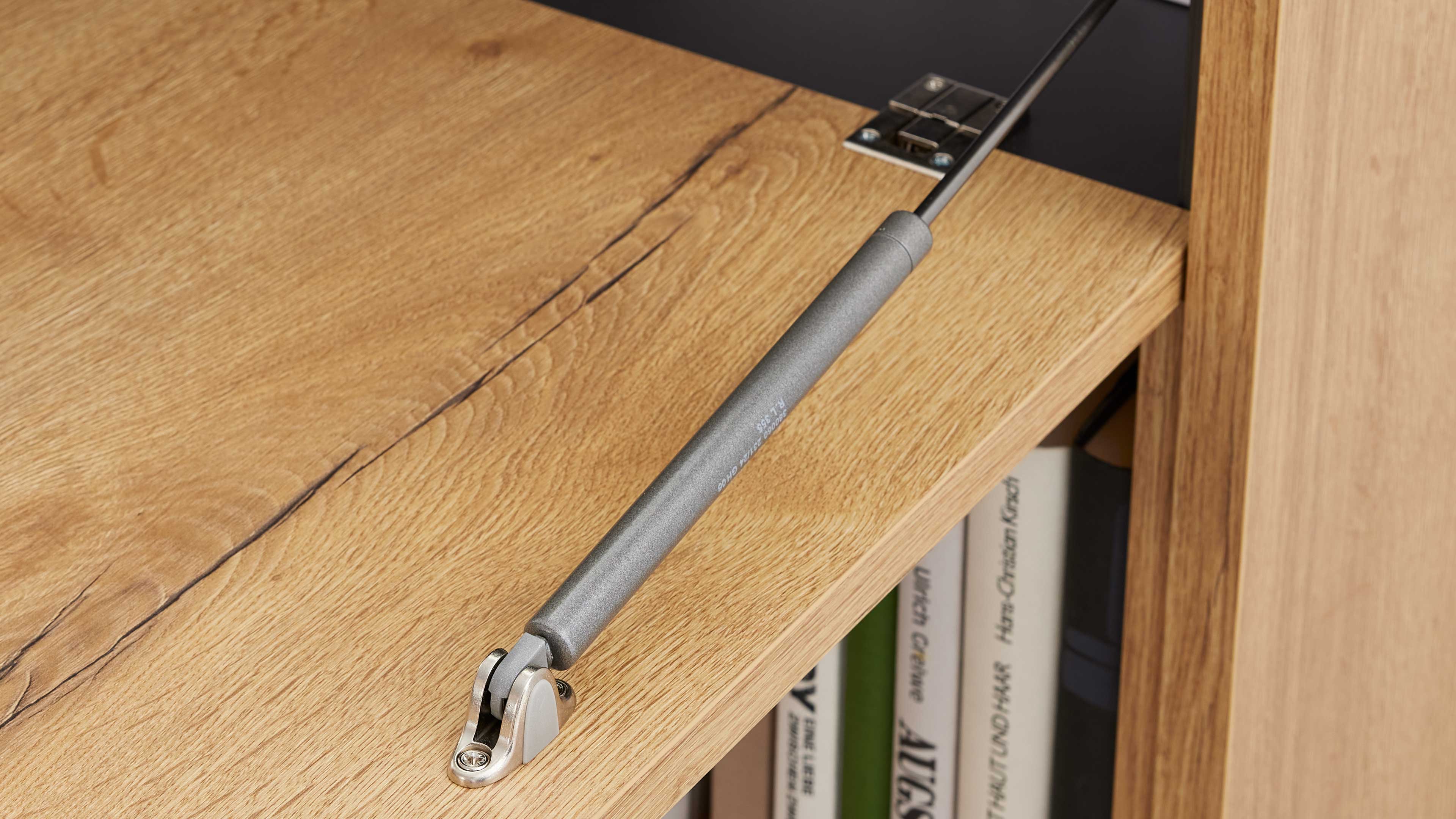 Eine silbergraue Gasdruckfeder verbindet die Klappe mit dem Holzregal und ist am Regalboden sowie an einem Scharnier befestigt.