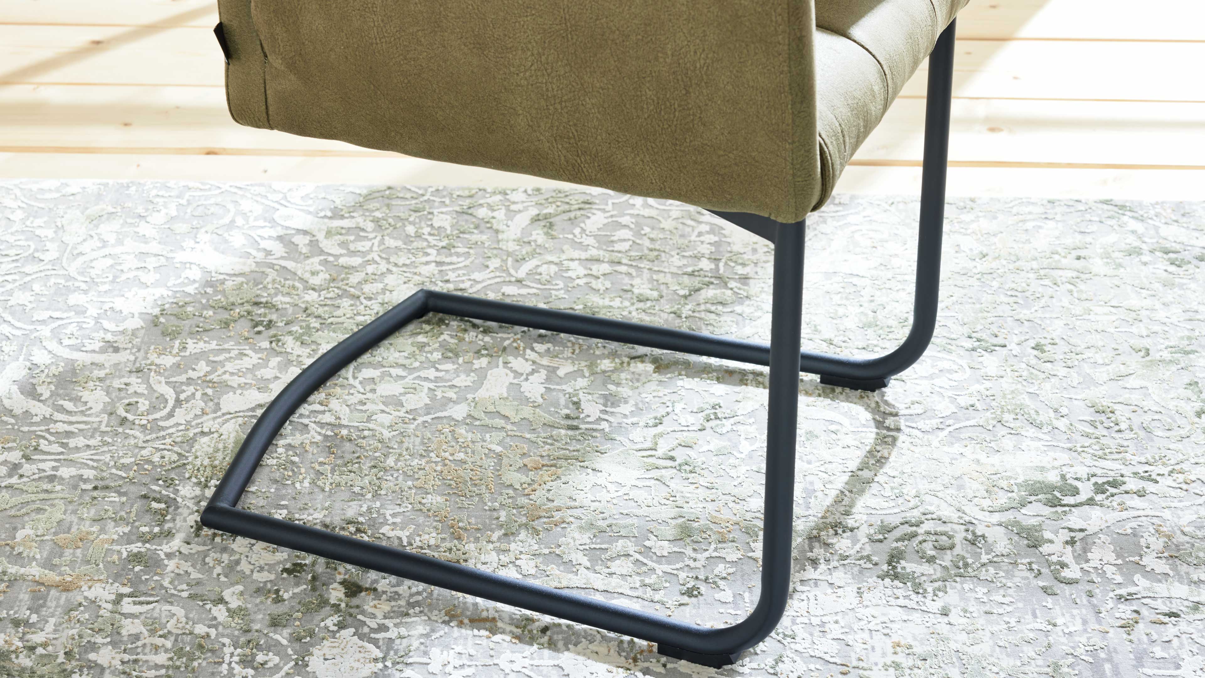 Unterer Teil eines olivgrünen Polsterstuhls mit schwarzem Freischwinger-Gestell auf hellgemustertem Teppich vor Holzdielen.