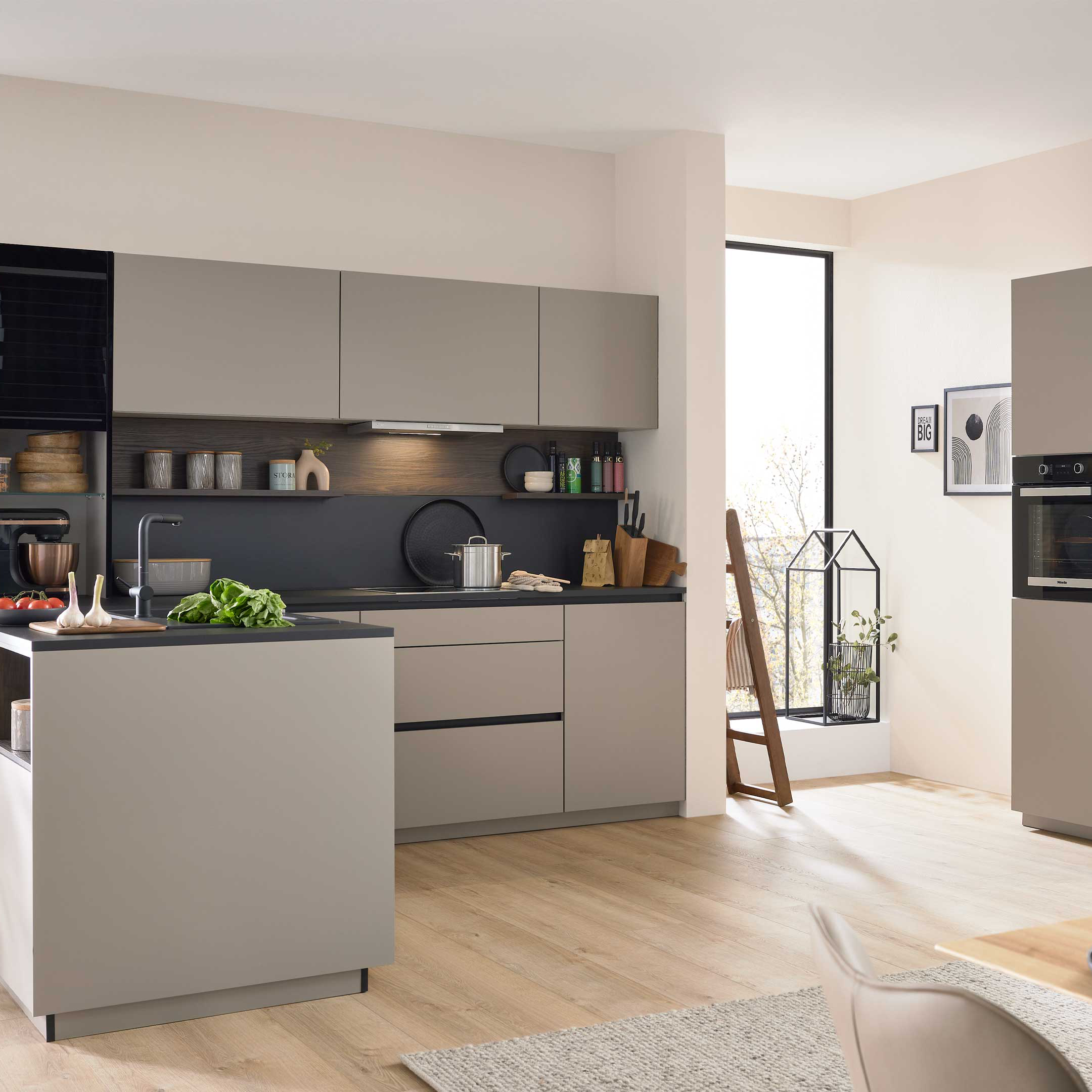 Moderne Küche mit hellgrauer Küchenzeile und Kochinsel, schwarzer Arbeitsplatte, Hochschrank mit Backofen und Pflanzen.