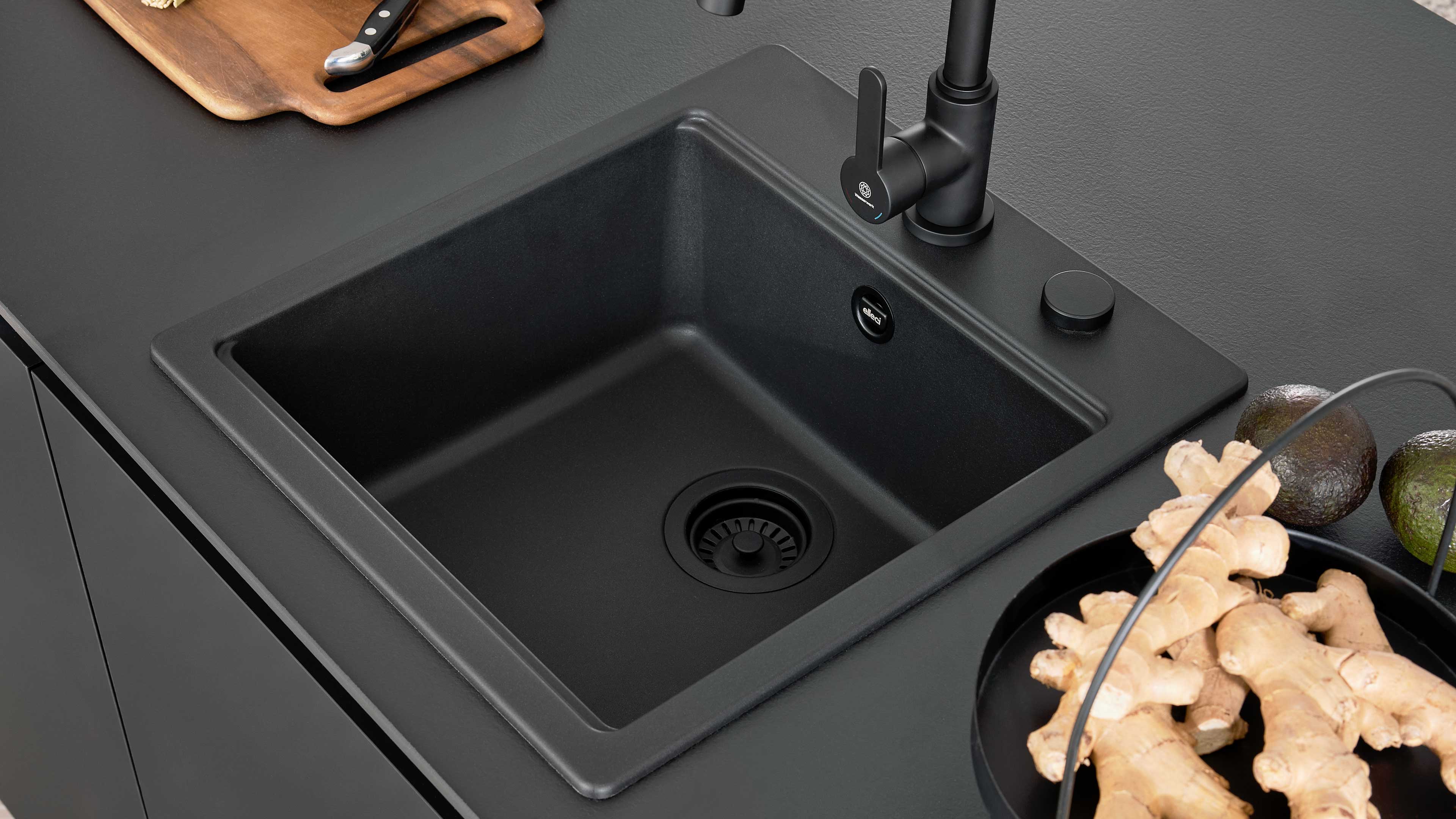 Schwarze eckige Küchenspüle mit schwarzem Einhebelmischer in dunkler Arbeitsplatte, daneben Schneidebrett, Ingwer und Avocados.