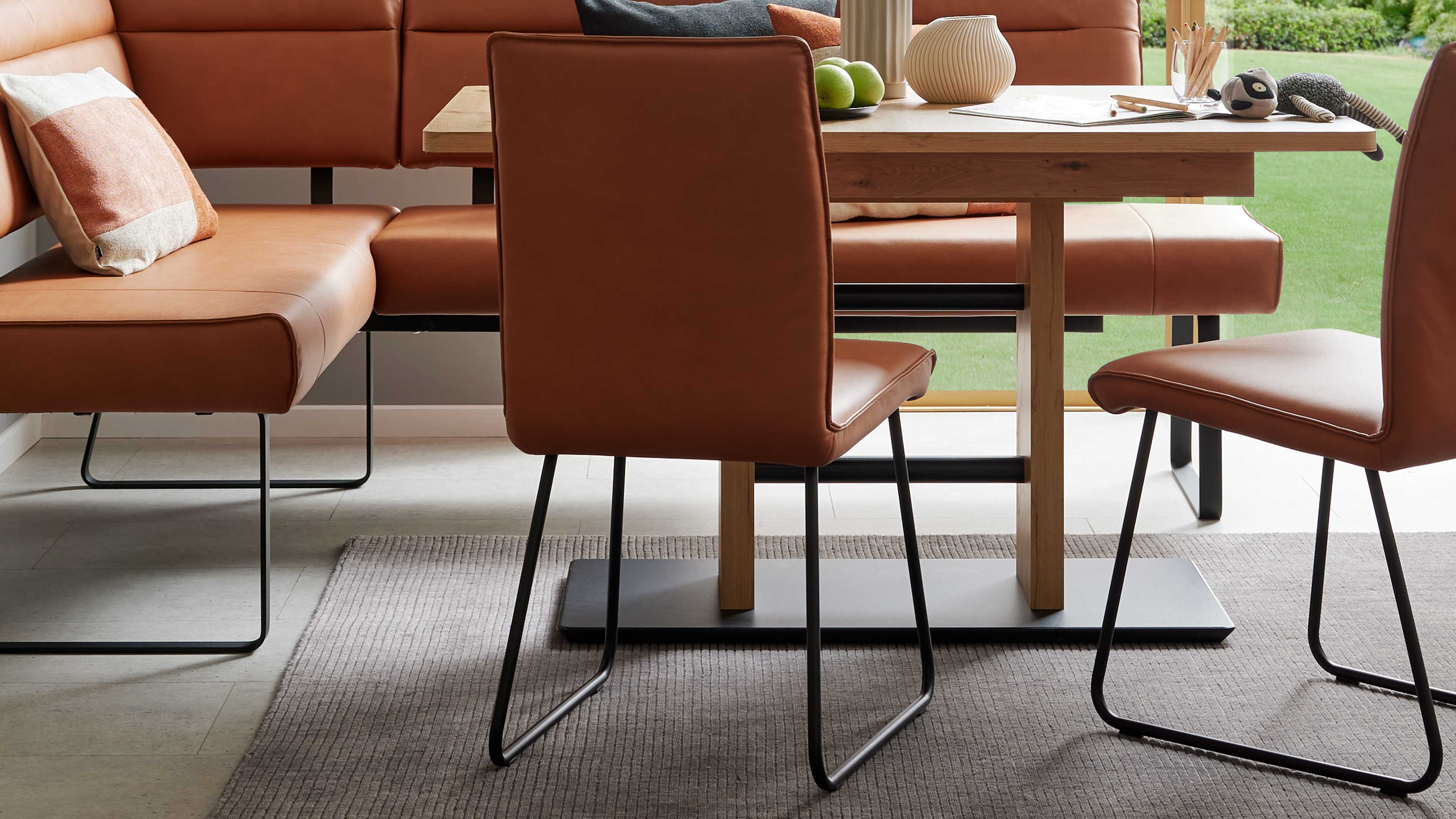 Braun gepolsterter Stuhl mit schwarzen Kufen vor einem Eichentisch, daneben Eckbank, Teppich und Tischdekoration.