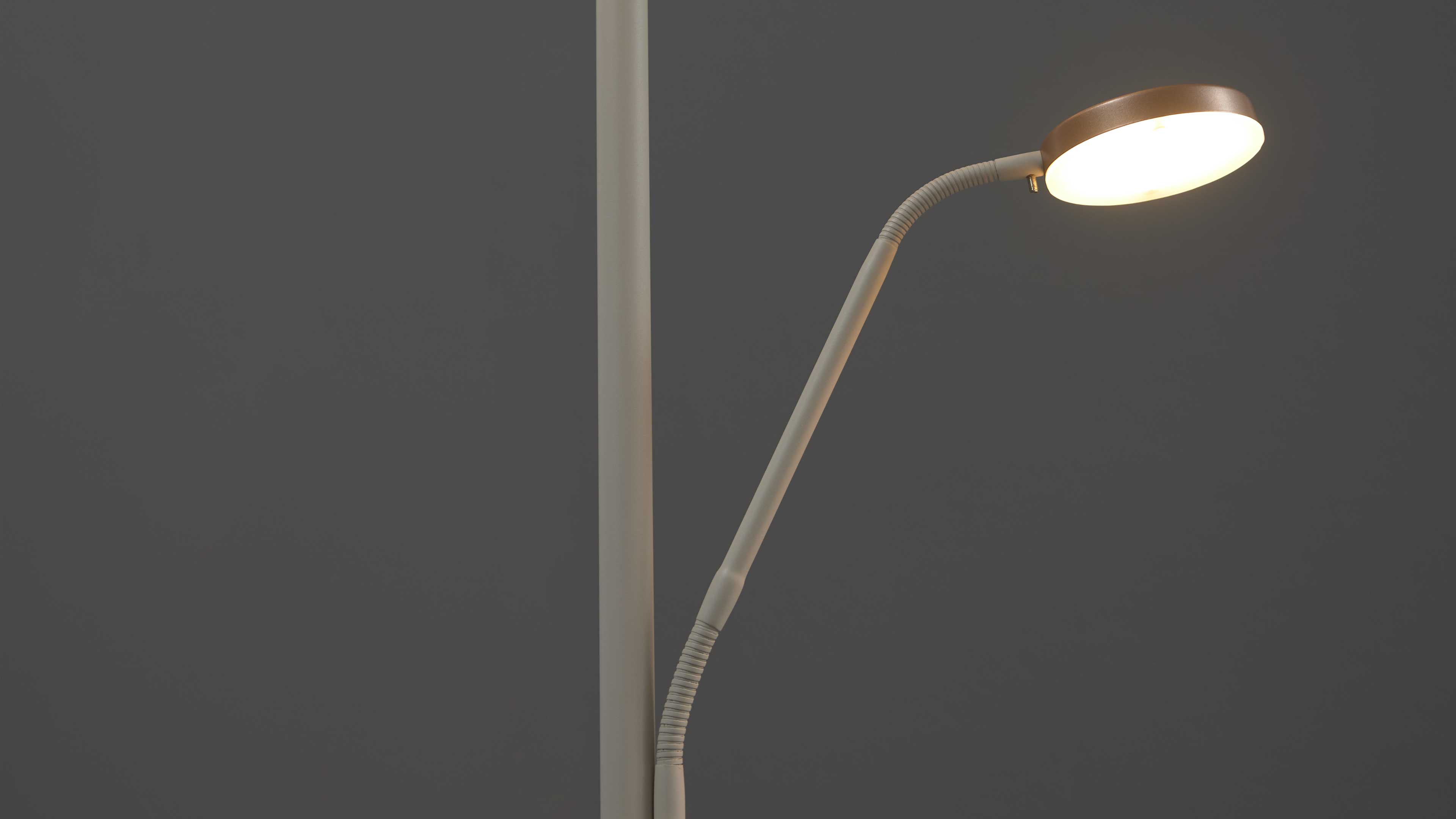 Stehlampe mit Ständer, flexiblem Gelenkarm und rundem, beleuchtetem Leuchtkopf mit bronzefarbenem Rand vor dunkelgrauem Hintergrund.