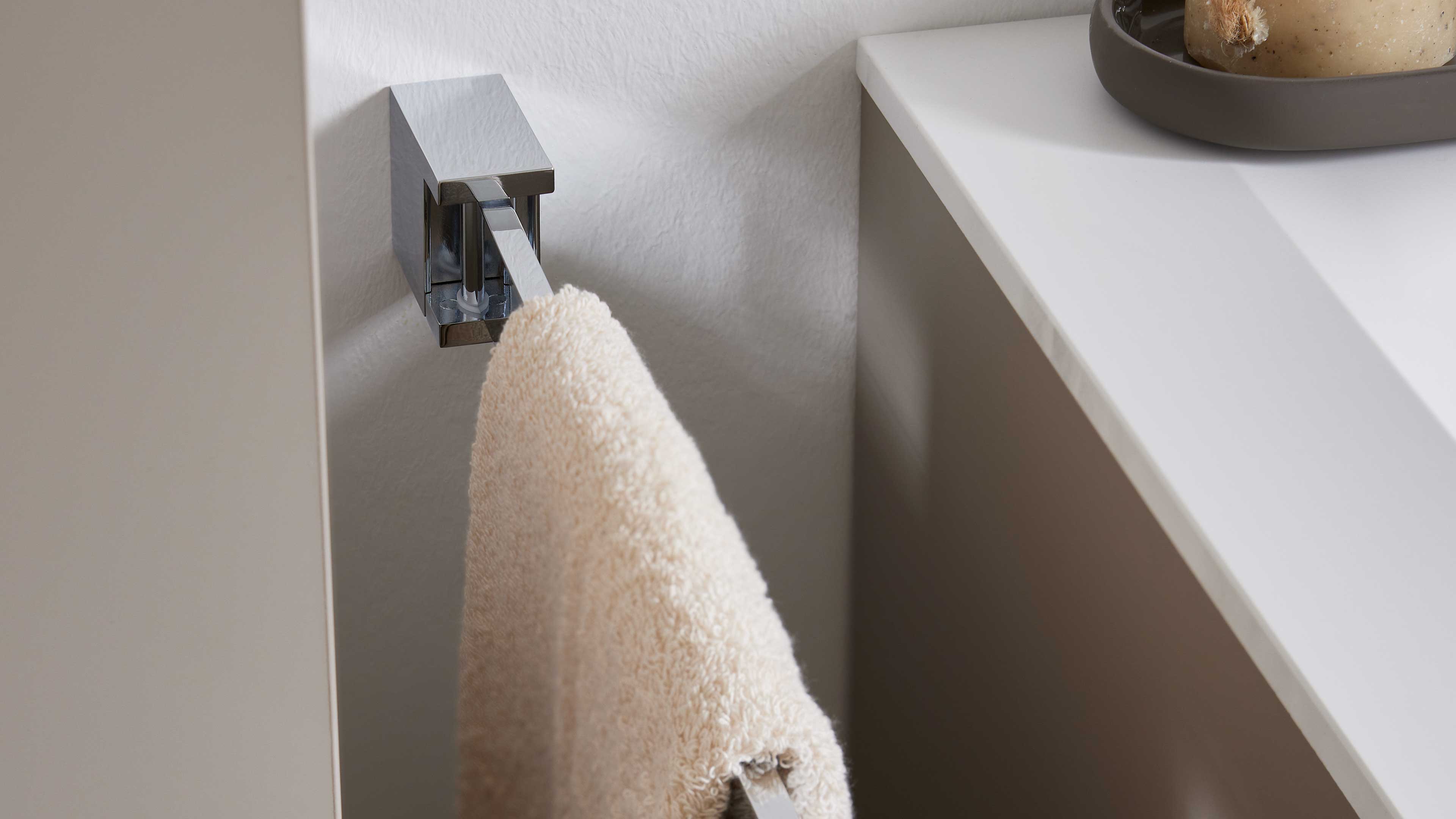 Beiges Handtuch hängt an einem chromglänzenden, eckigen Wand-Handtuchhalter neben einer weißen Waschtischplatte.
