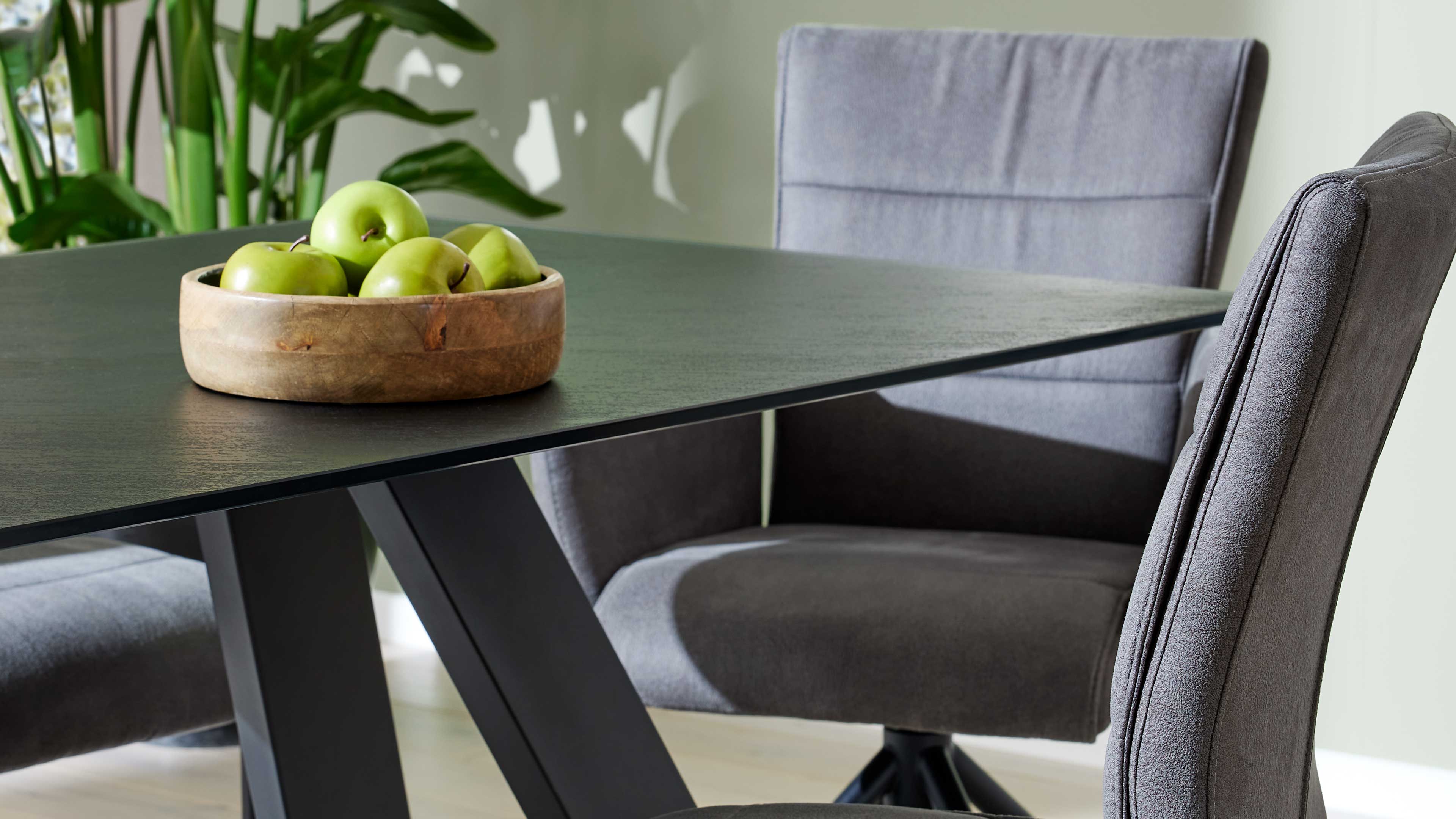 Schwarzer Esstisch mit runder Holzschale voller grüner Äpfel, dazu graue, gepolsterte Stühle und grüne Zimmerpflanze.