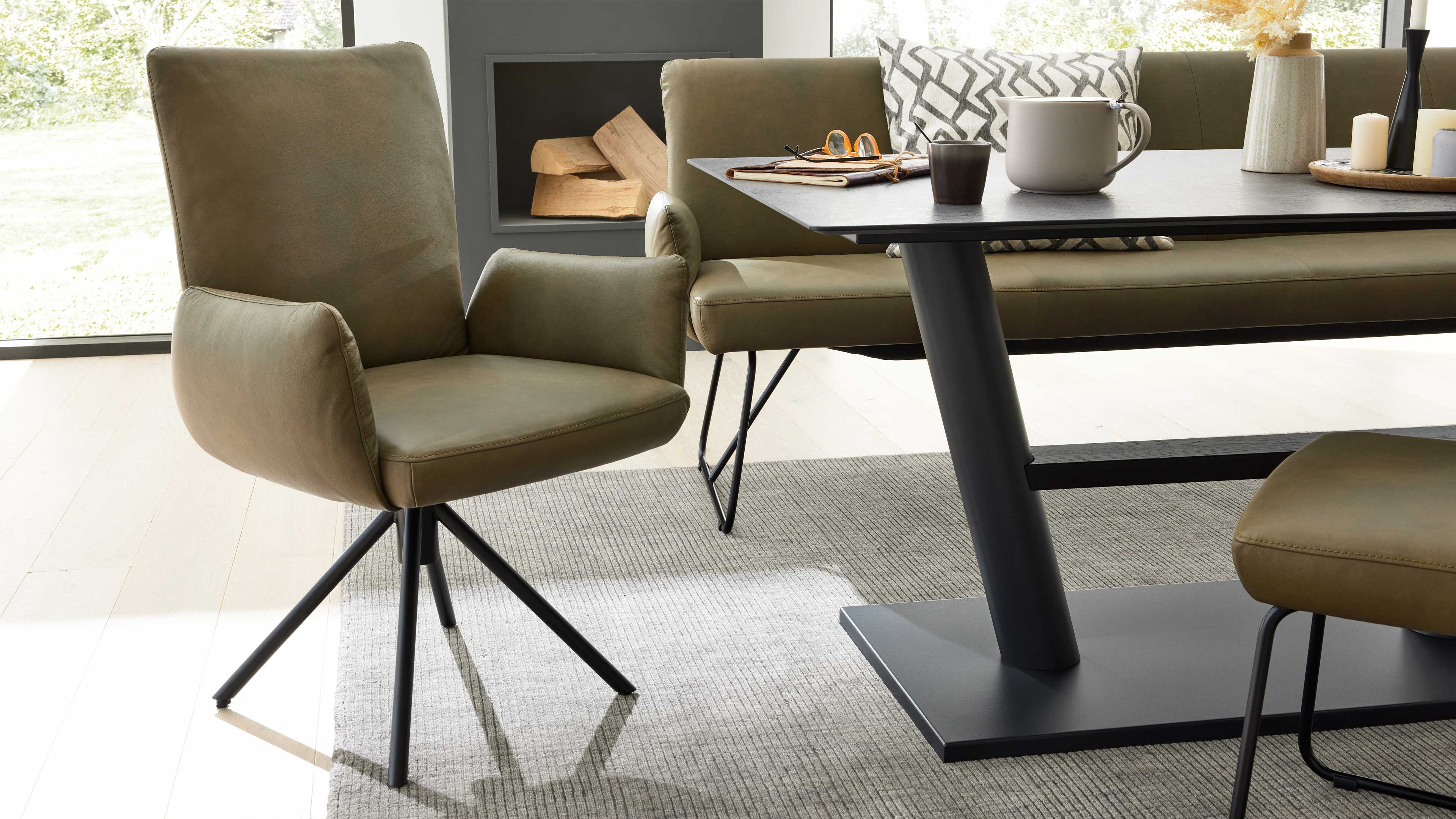 Olivegrüner, gepolsterter Schwenksessel mit schwarzem Metallsternfuß, daneben Tisch, Bank, Teppich und Kerzen.